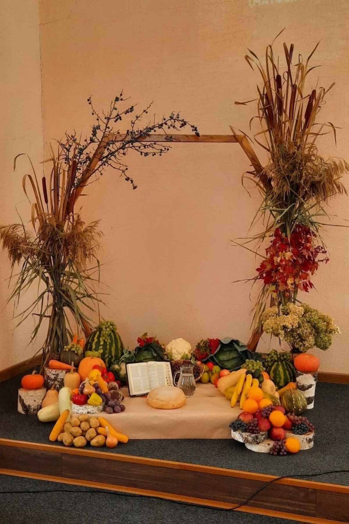 A harvest display featuring an open book and loaf of bread surrounded by various fruits and vegetables, framed by a wooden arch decorated with dried plants.