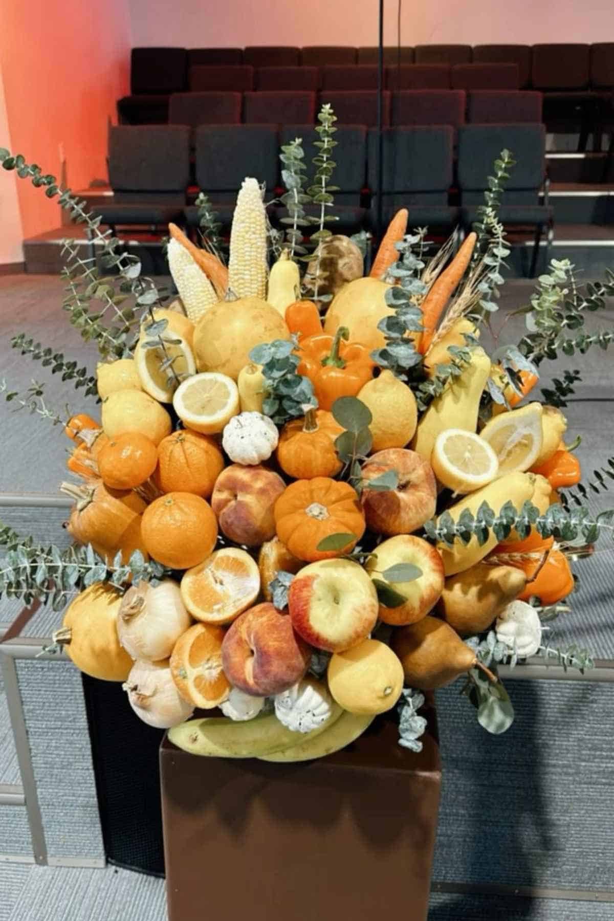 A large, overflowing arrangement of assorted fresh fruits, vegetables, and eucalyptus stems in a display stand.