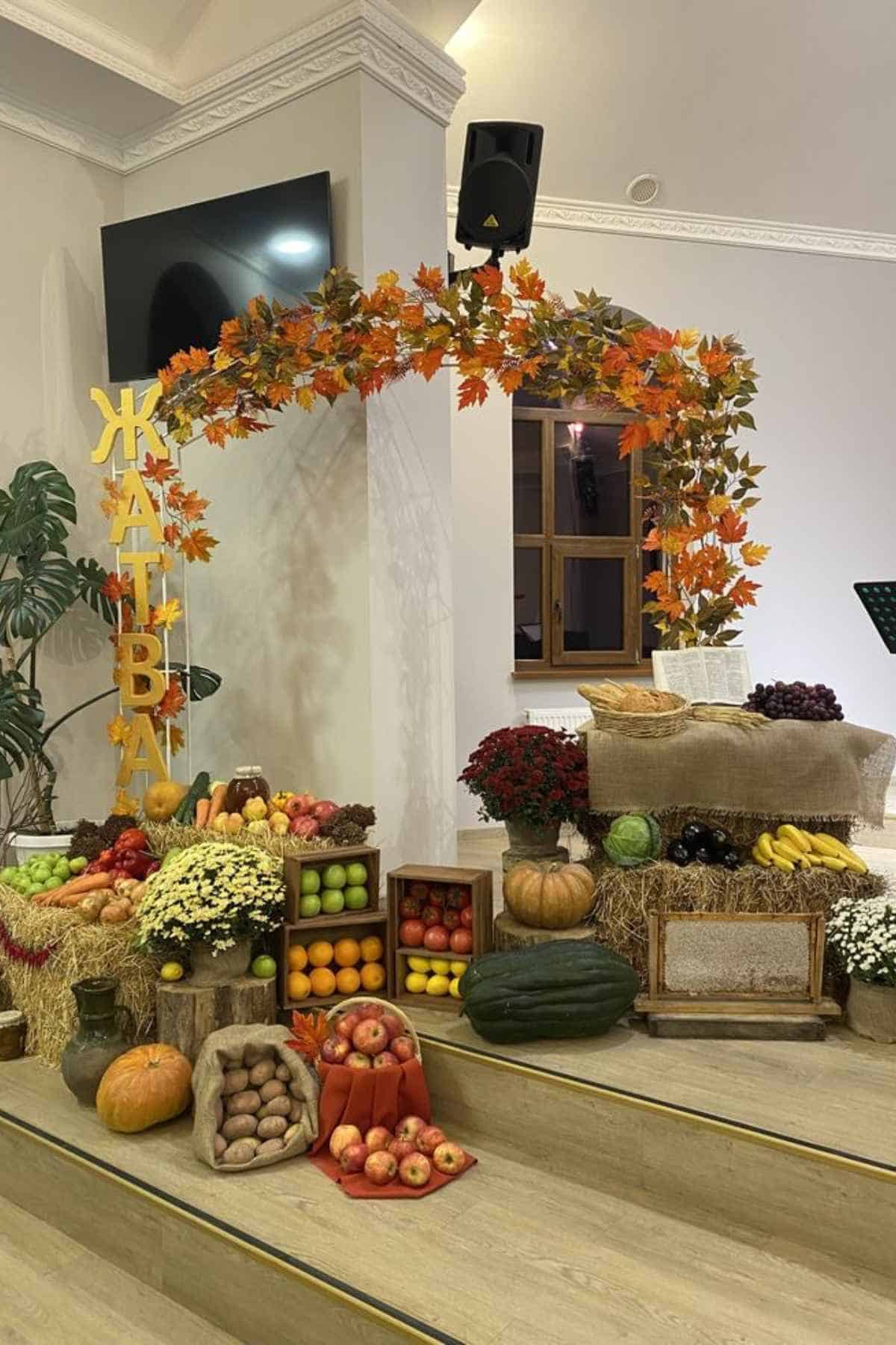 An autumn harvest display featuring wooden crates and bales of hay filled with apples, pumpkins, gourds, and other produce, decorated with an arched branch of orange maple leaves and the letters “ЖАТВА.”