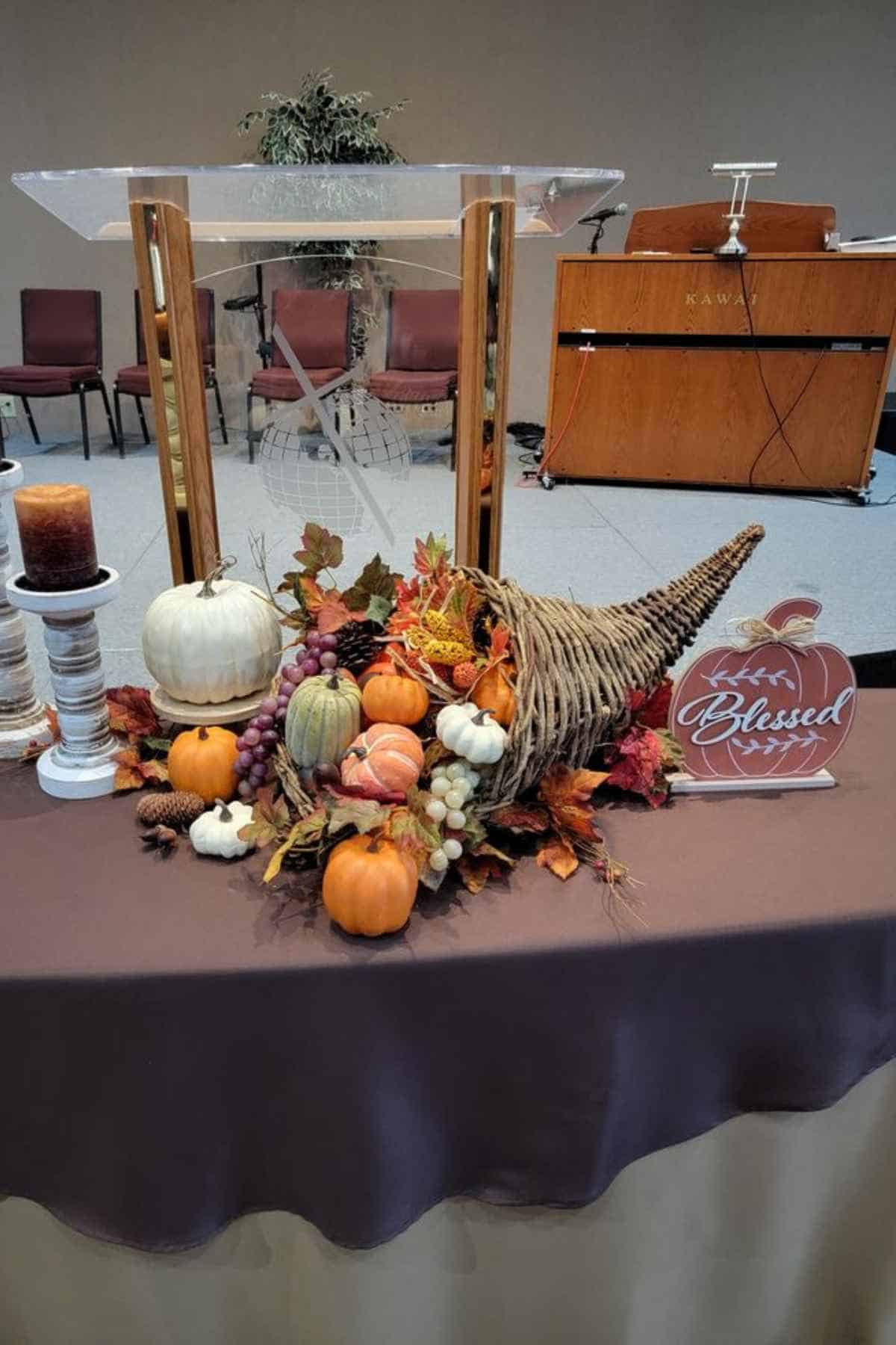 A cornucopia filled with small pumpkins, gourds, and artificial autumn leaves on a table with a wooden “Blessed” sign and a candle.