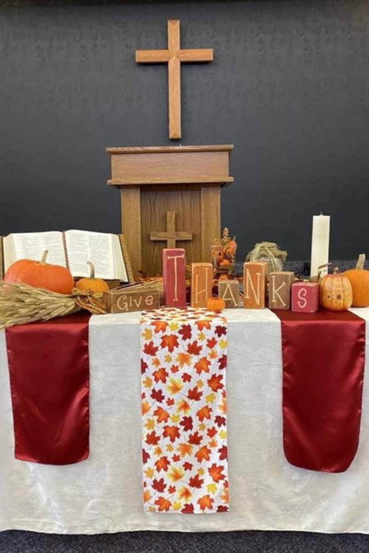 An altar decorated for Thanksgiving with an open Bible, pumpkins, and wooden blocks spelling “Give Thanks” in front of a wooden cross.