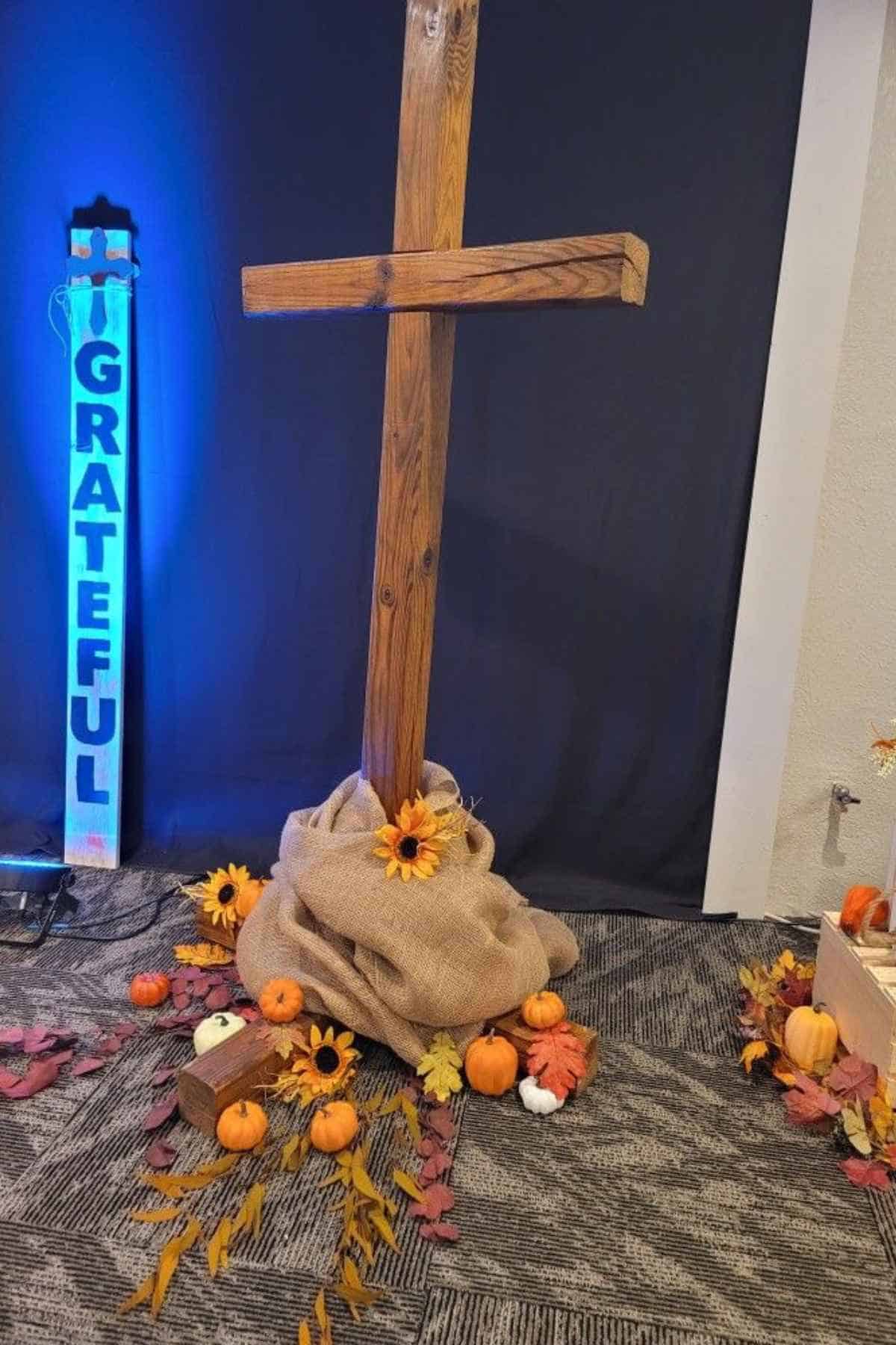 A wooden cross set in a burlap base on the floor, surrounded by small pumpkins and autumn leaves, next to a sign that reads “Grateful.”