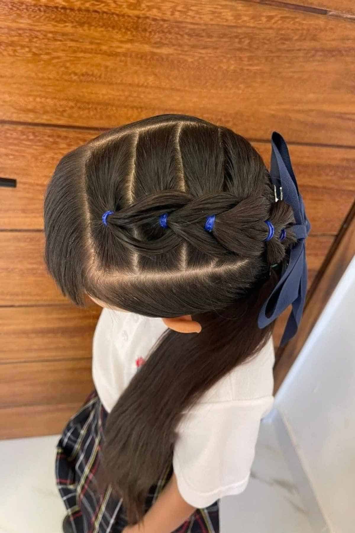 A child’s hair styled in a segmented French braid with blue elastic bands and a large blue hair bow.