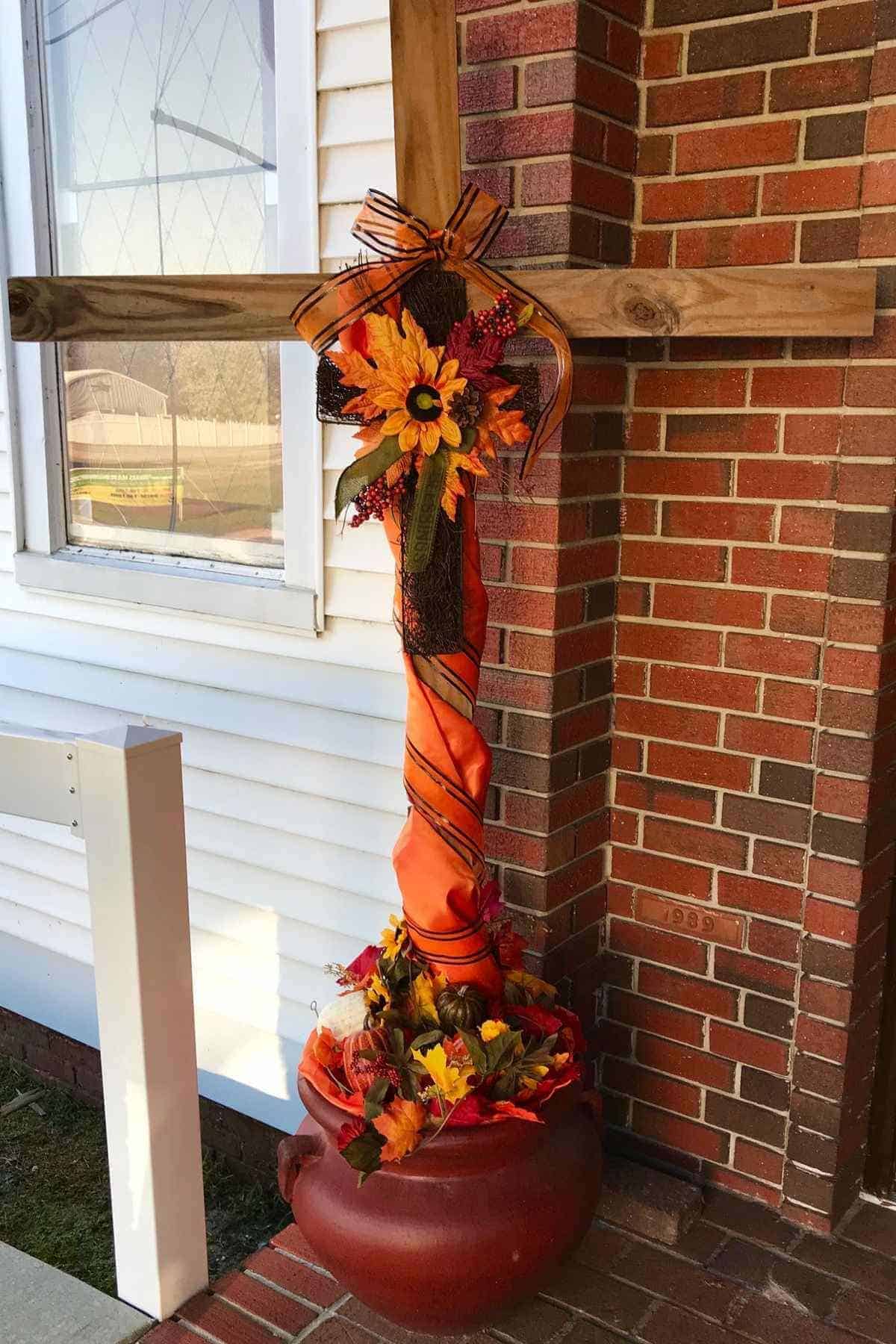 A wooden cross decorated with orange fabric, autumn leaves, a sunflower, and berries, standing in a large pot filled with seasonal foliage.