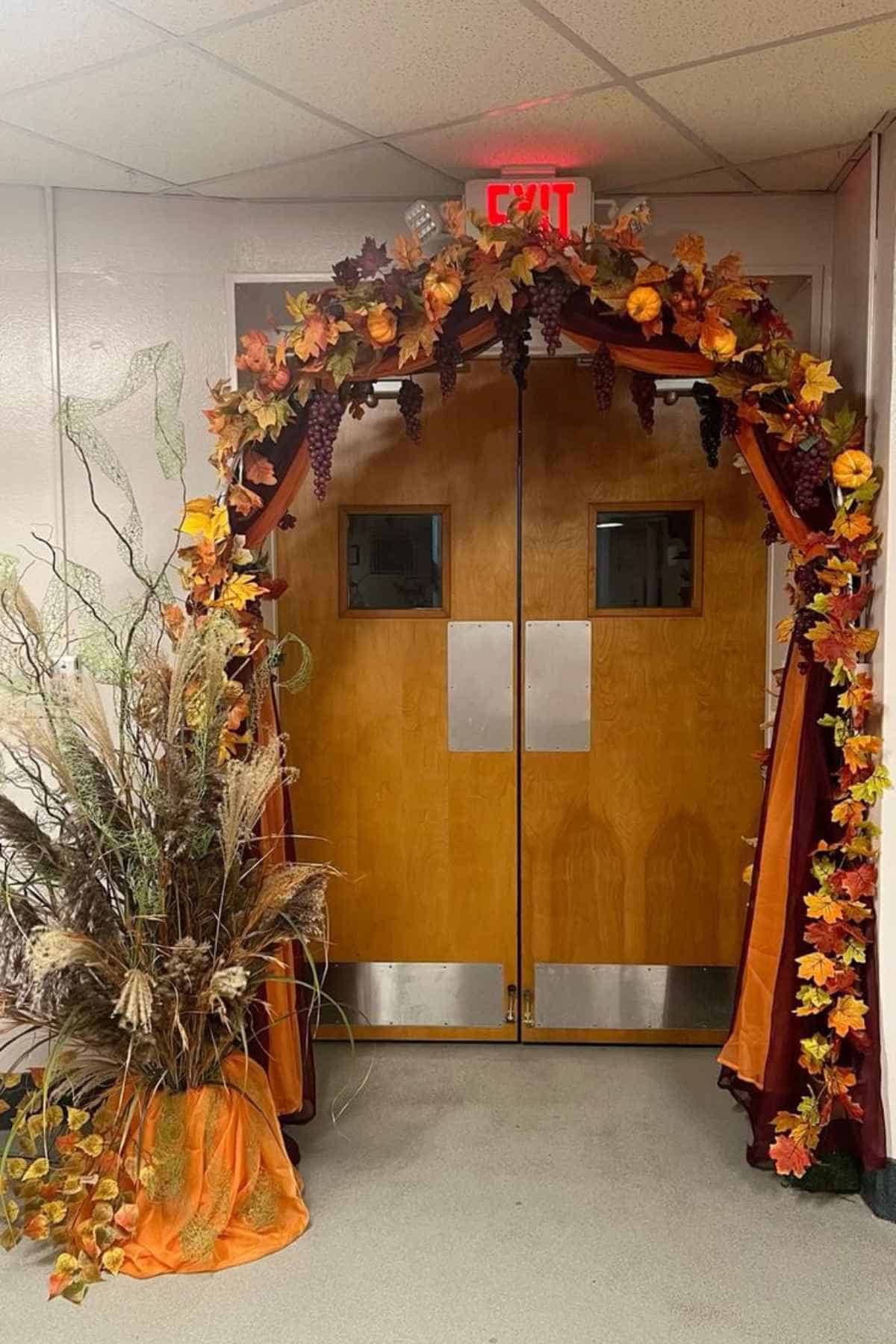 Double wooden doors decorated with an autumnal arch of fall leaves, pumpkins, and grapes, with a dried floral arrangement to the left.
