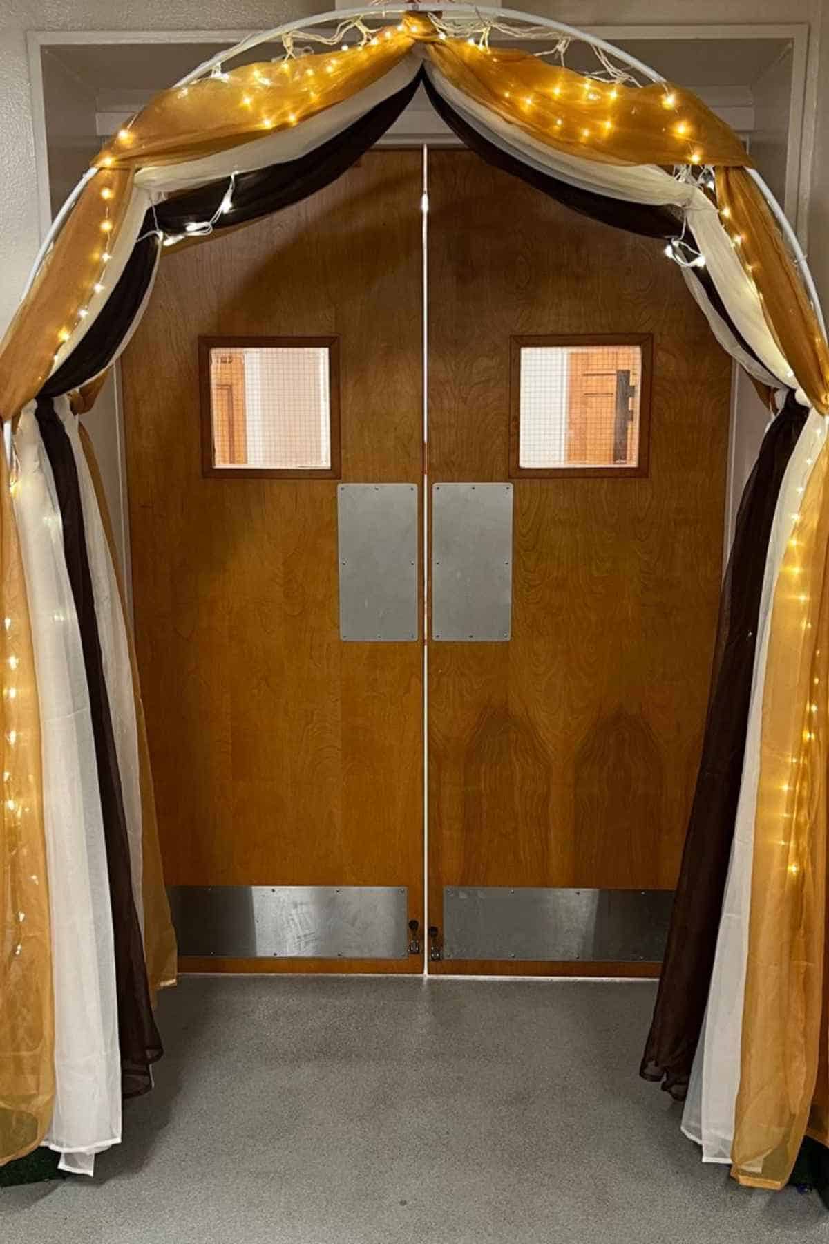 Double wooden doors framed by a white arch draped in gold, brown, and white fabric with string lights.