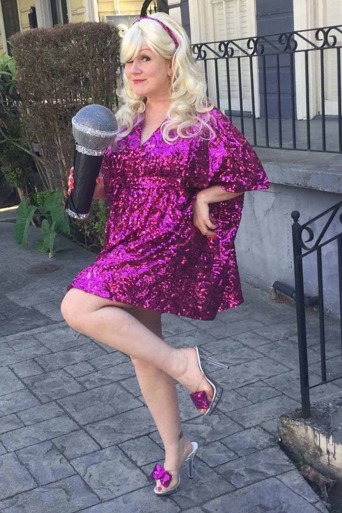 Person in a shiny pink sequined dress, blonde wig, and silver heels poses outdoors holding a large, glittery microphone prop.