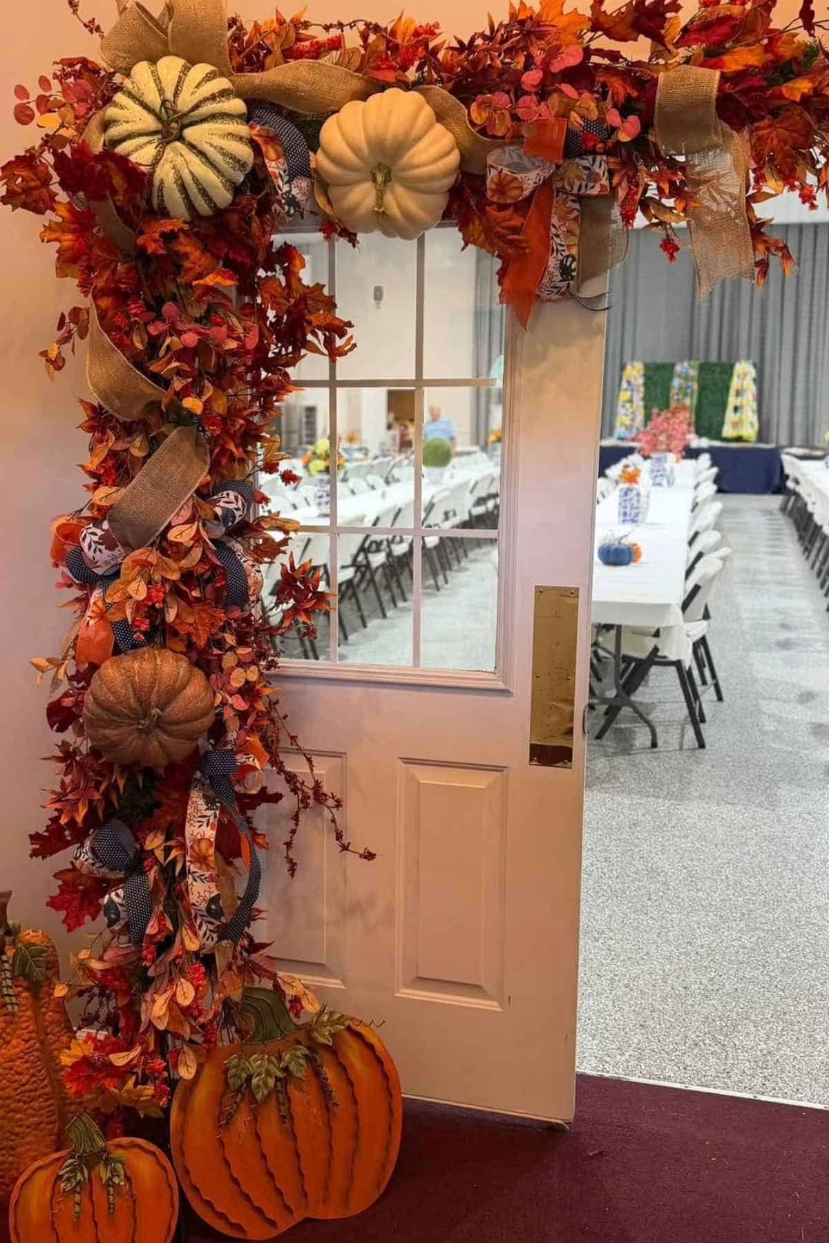A doorway decorated with autumn foliage, ribbons, and decorative pumpkins, opening into a hall with rows of tables set for an event.