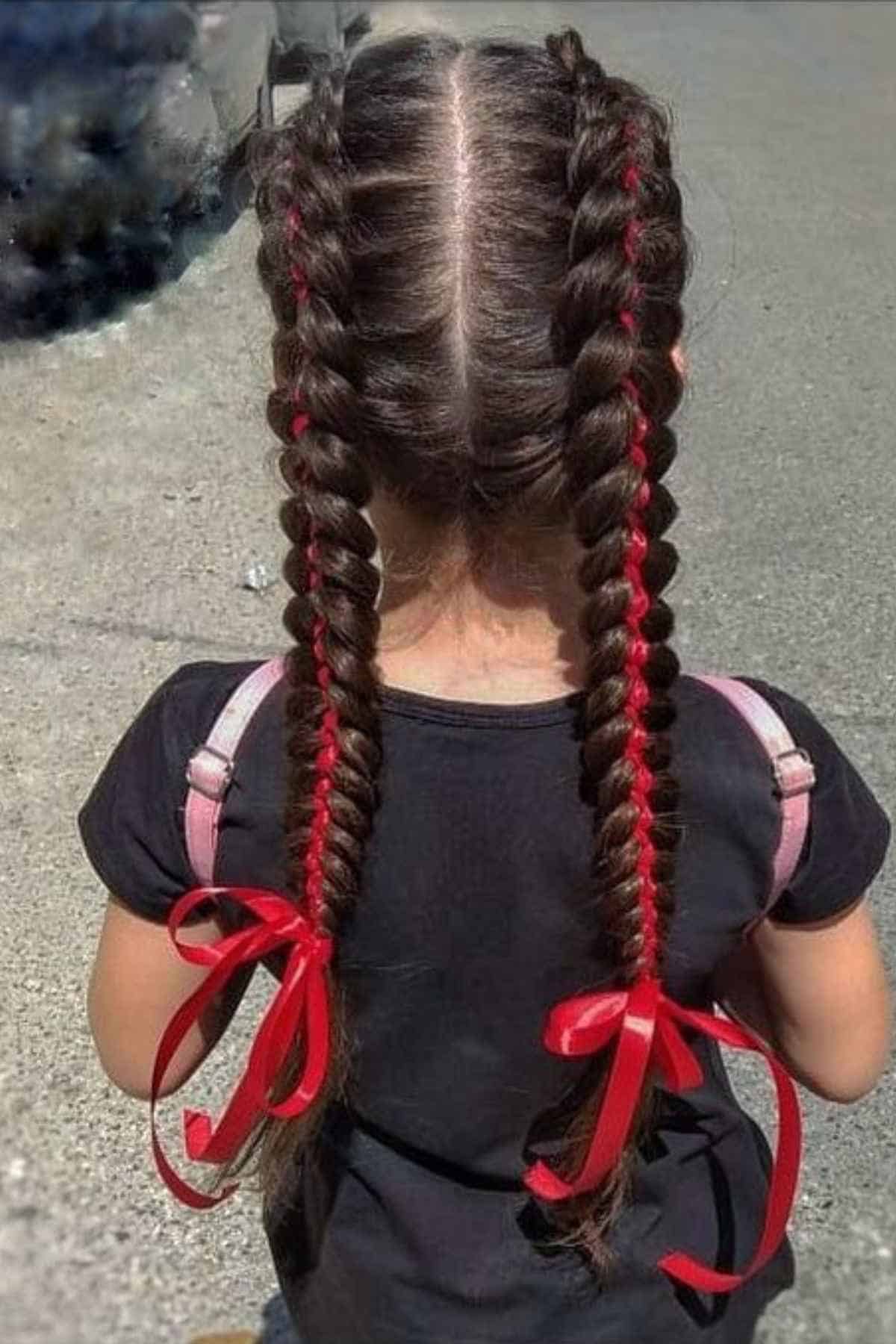 A child seen from behind with two French braids styled with red ribbon threaded through the hair and tied in bows at the ends.