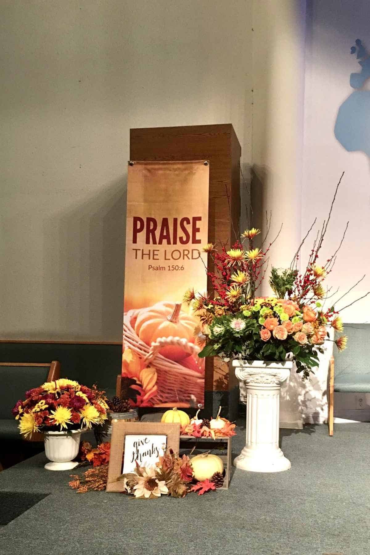 A banner reading “Praise the Lord, Psalm 150:6” with a pumpkin basket, a large floral arrangement on a pedestal, and a small “give thanks” sign on a church stage.