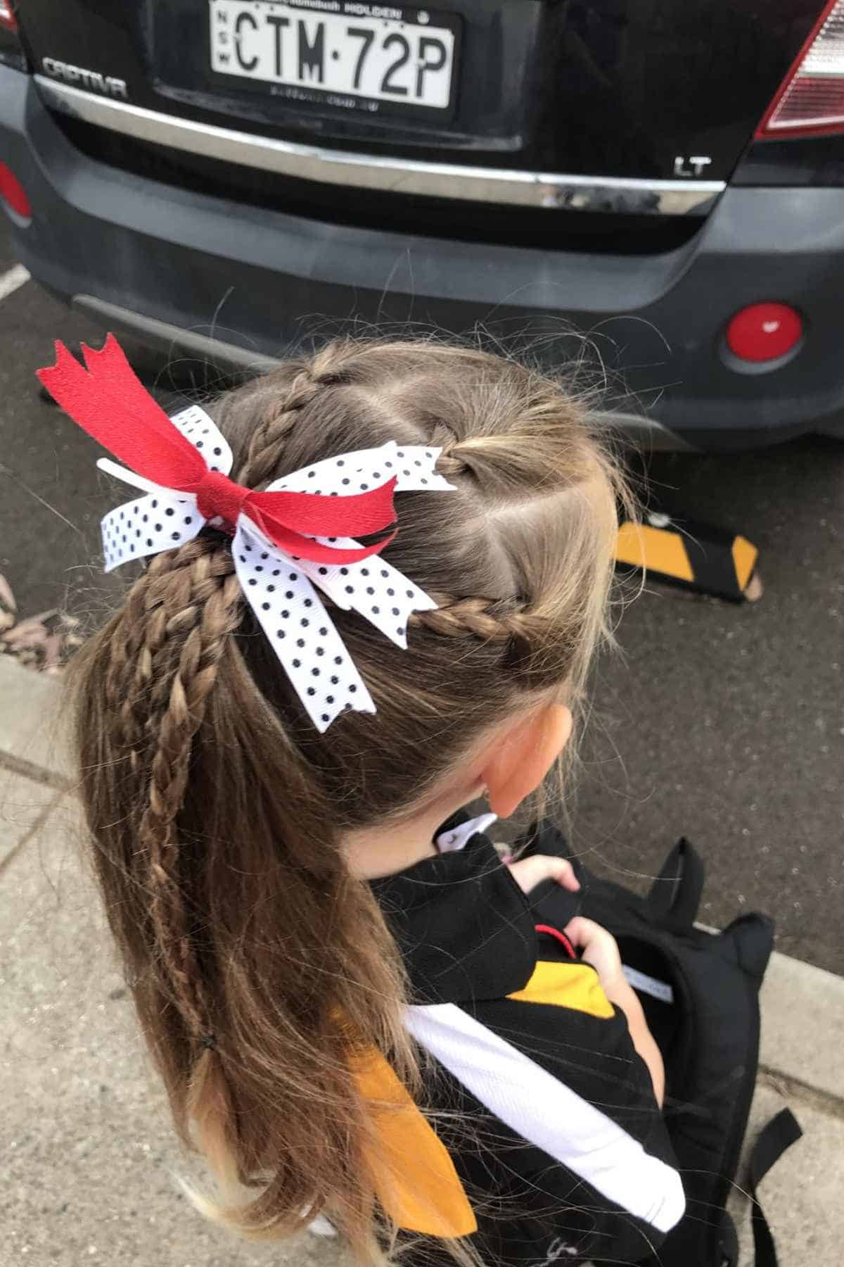 A child with braided hair tied in a ponytail with a red and white polka-dot bow, standing behind a car.
