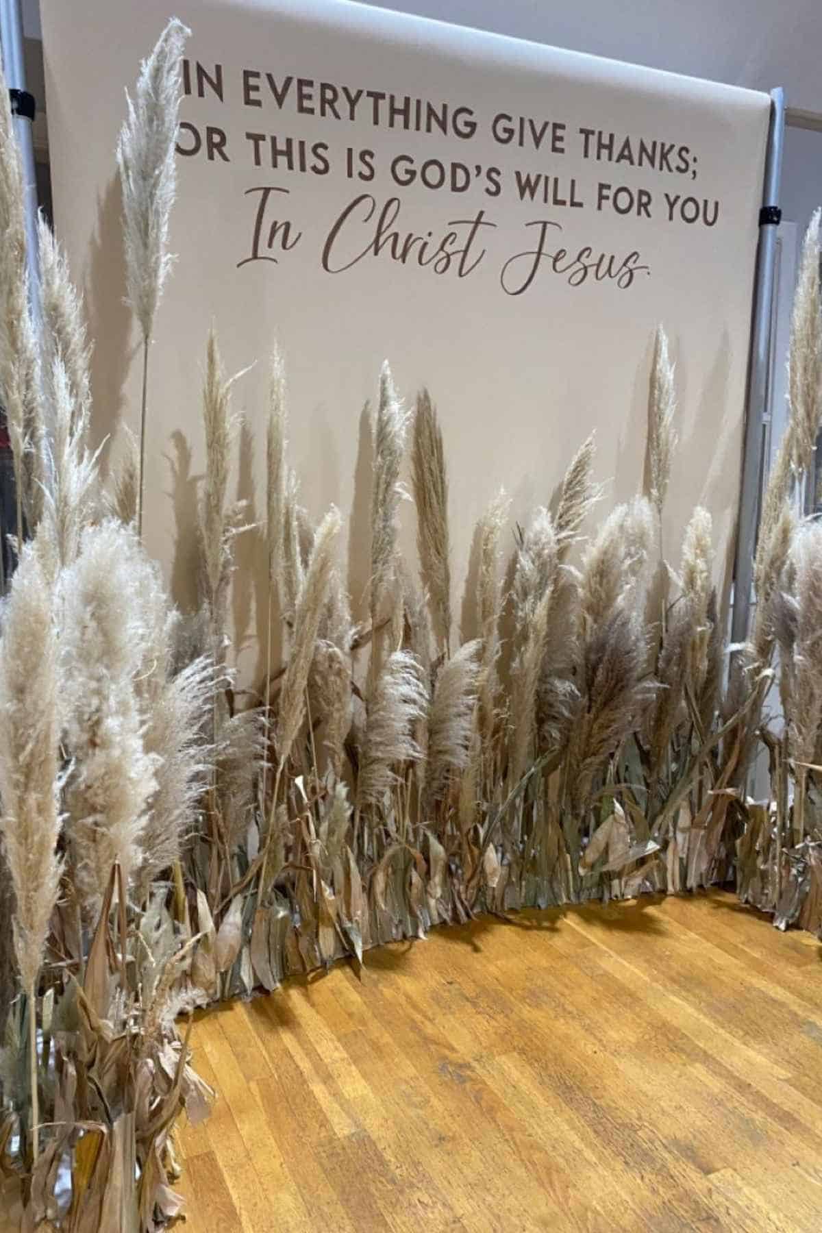A backdrop decorated with pampas grass that reads, “In everything give thanks; for this is God’s will for you In Christ Jesus.”