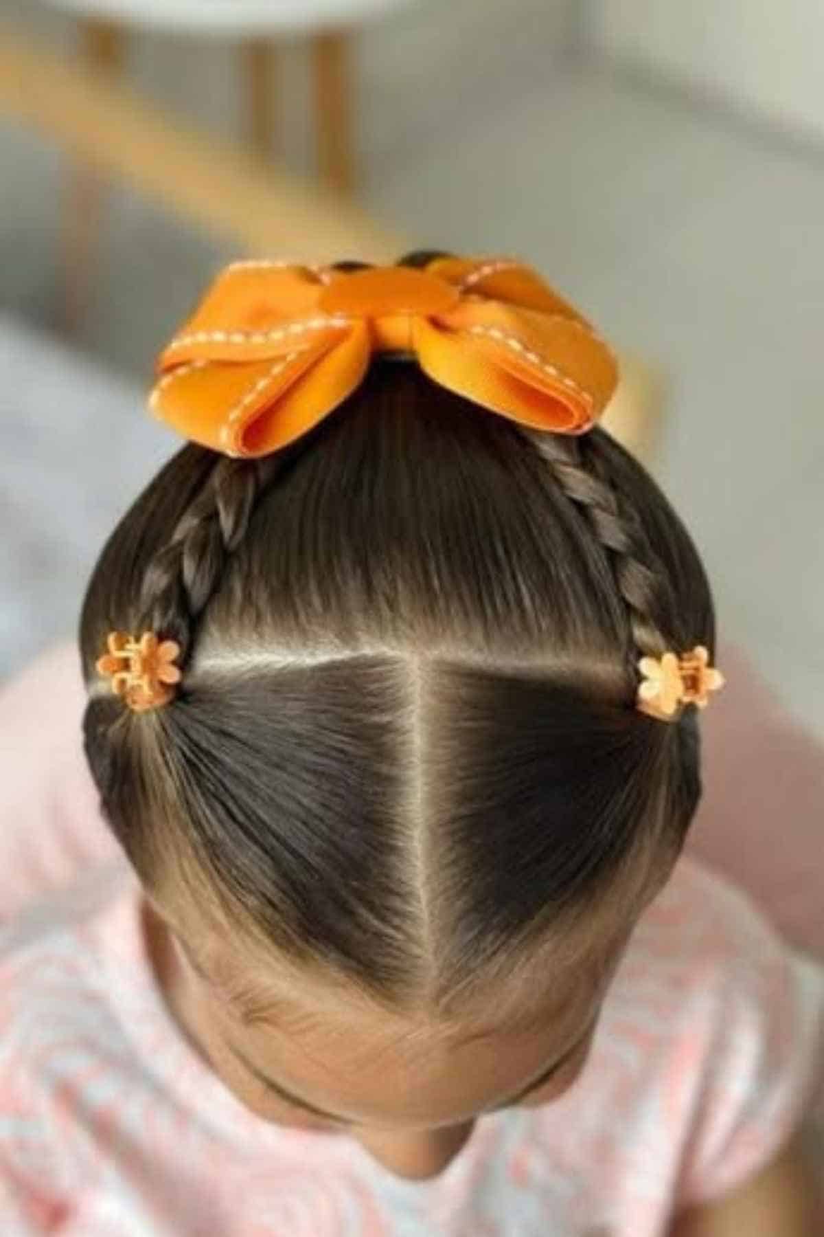 A child’s hair styled with two symmetrical braids leading into a high ponytail, accented with small orange flower clips and a large orange bow.
