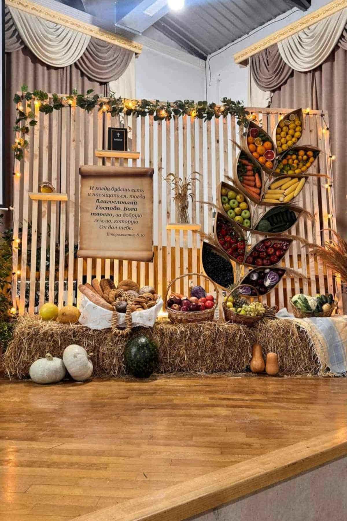 A wooden harvest display on straw bales, featuring baskets of fruit and vegetables, a scroll with Russian religious text, and a decorative leaf-shaped rack filled with produce.