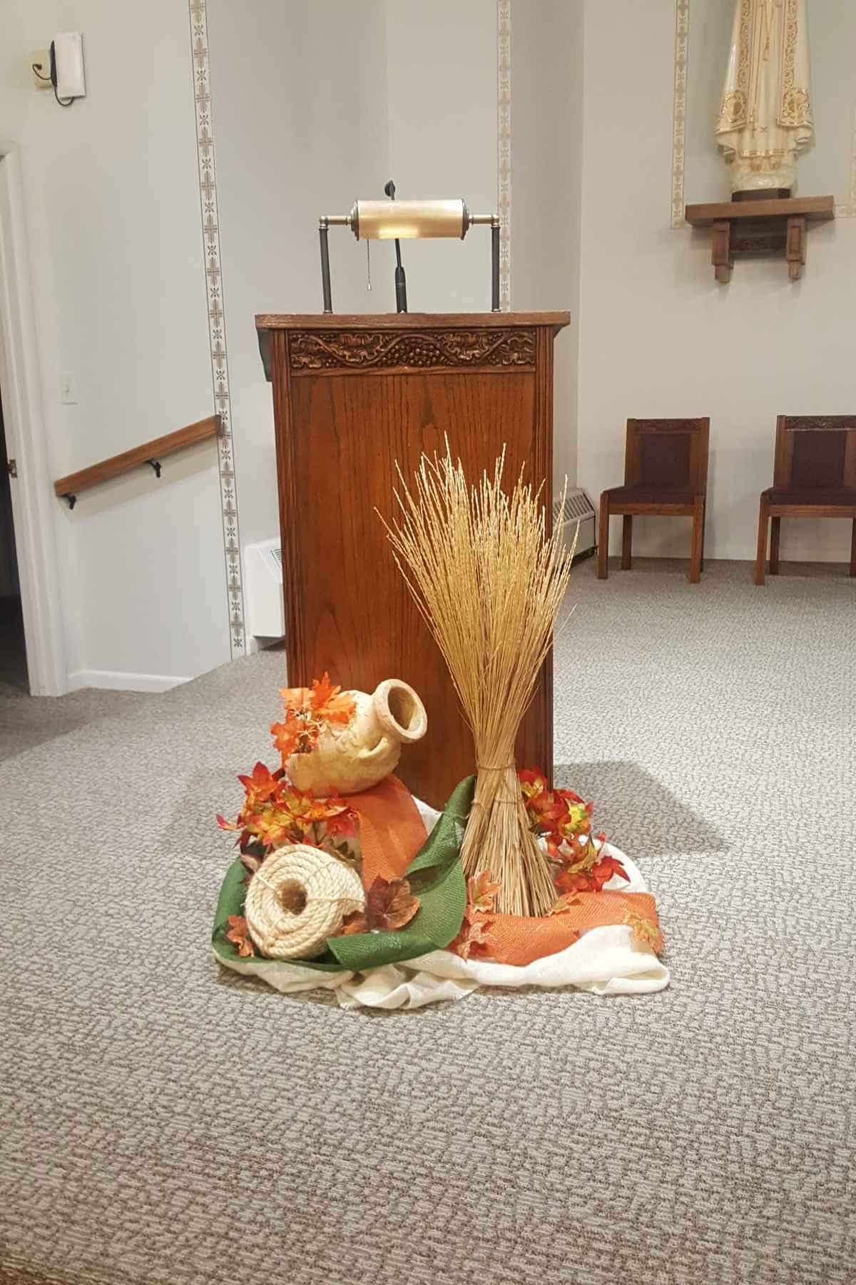 A wooden lectern decorated with a harvest display of wheat, a ceramic jug, and autumn leaves on the floor in front of it.