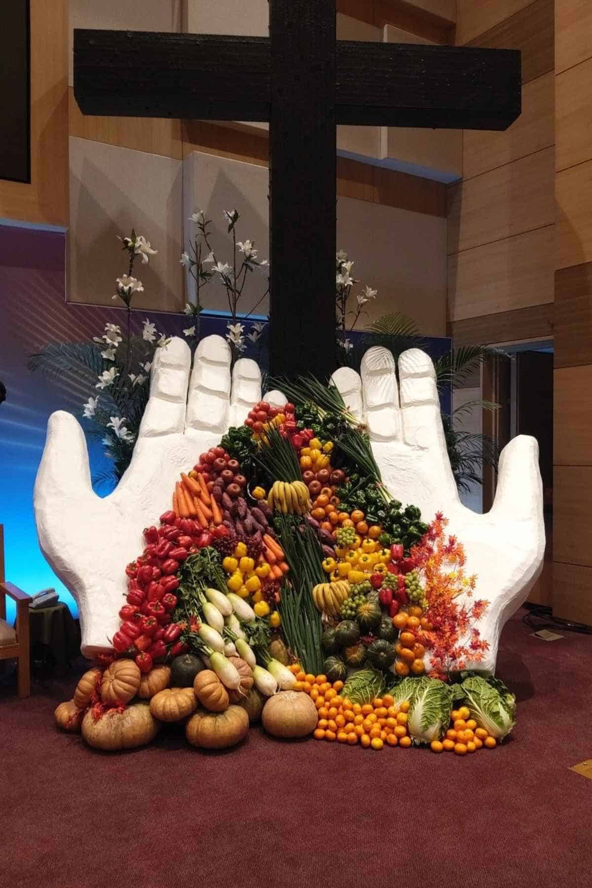 A large, white sculpture of two cupped hands overflowing with a variety of fresh produce, placed in front of a dark wooden cross.
