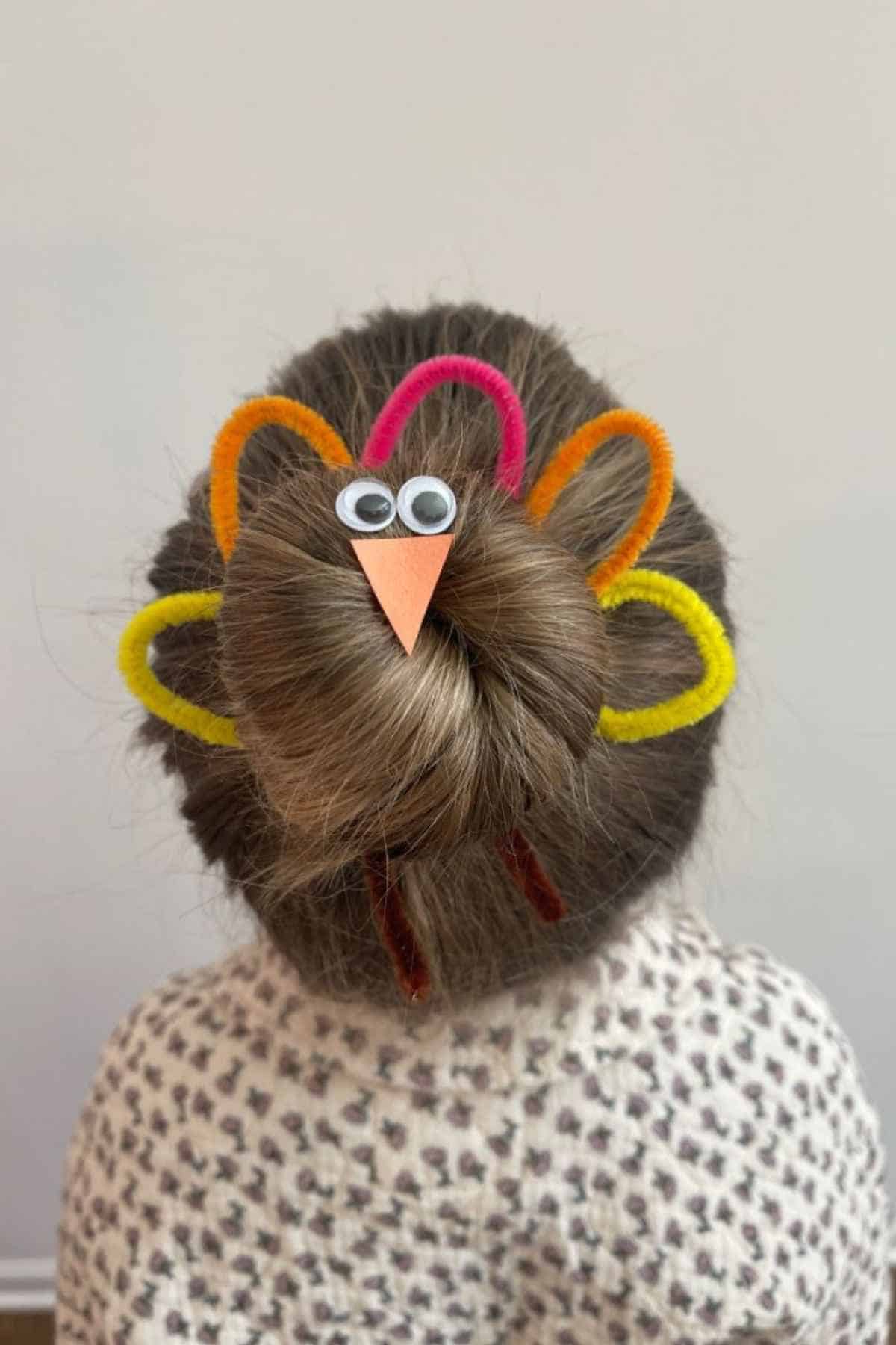 A hair bun styled to look like a turkey using googly eyes, a paper beak, and colorful pipe cleaners.