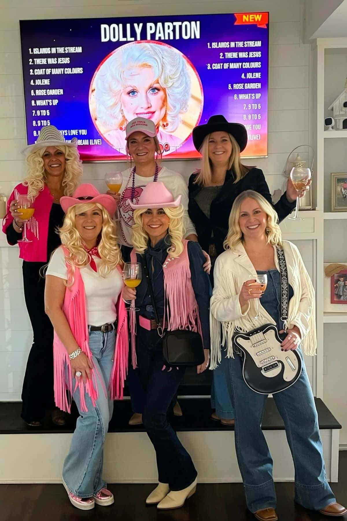 Six women in country-themed costumes pose indoors holding drinks, with a Dolly Parton playlist displayed on a TV screen behind them.