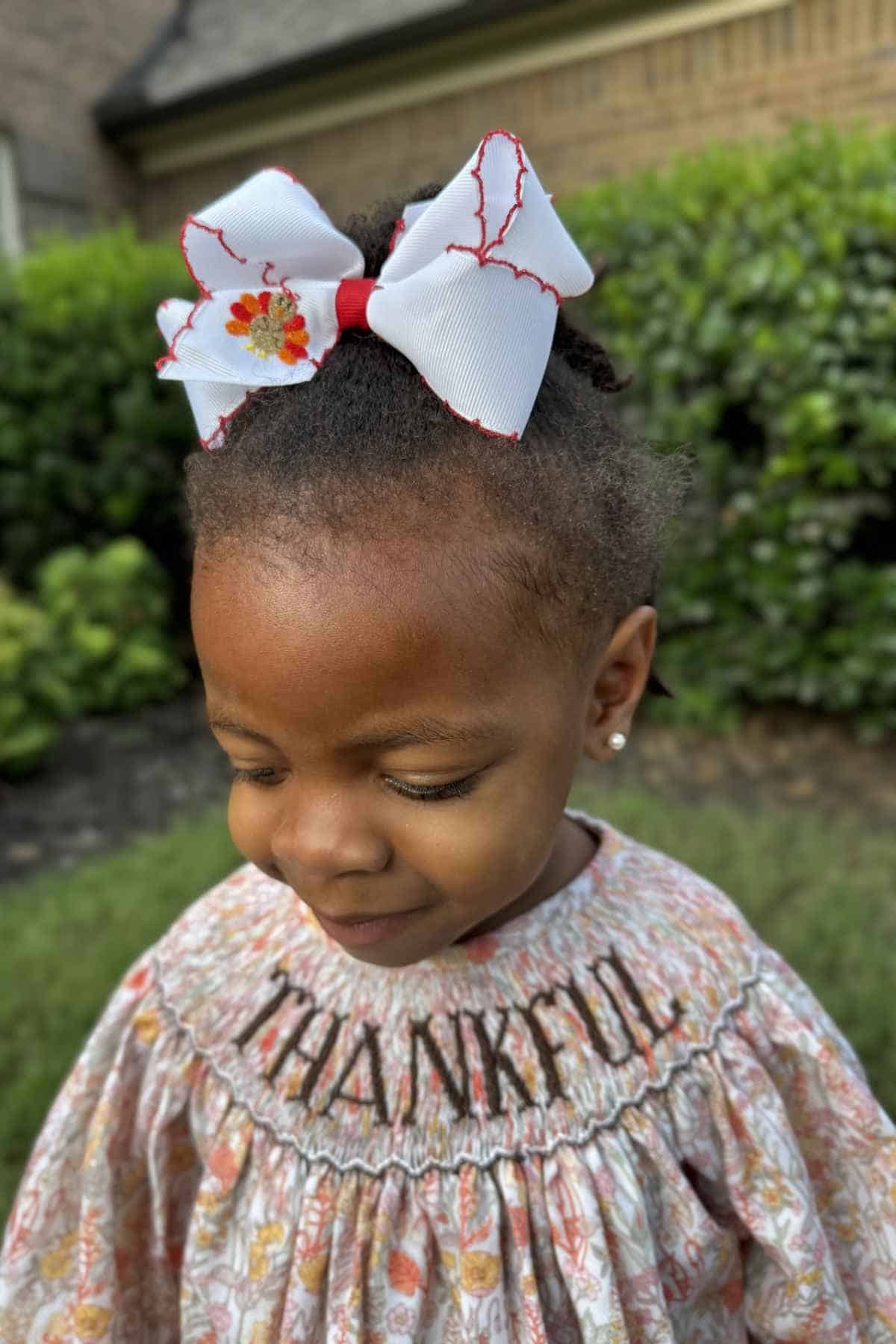 A young child wearing a floral dress embroidered with the word “THANKFUL” and a large white hair bow.