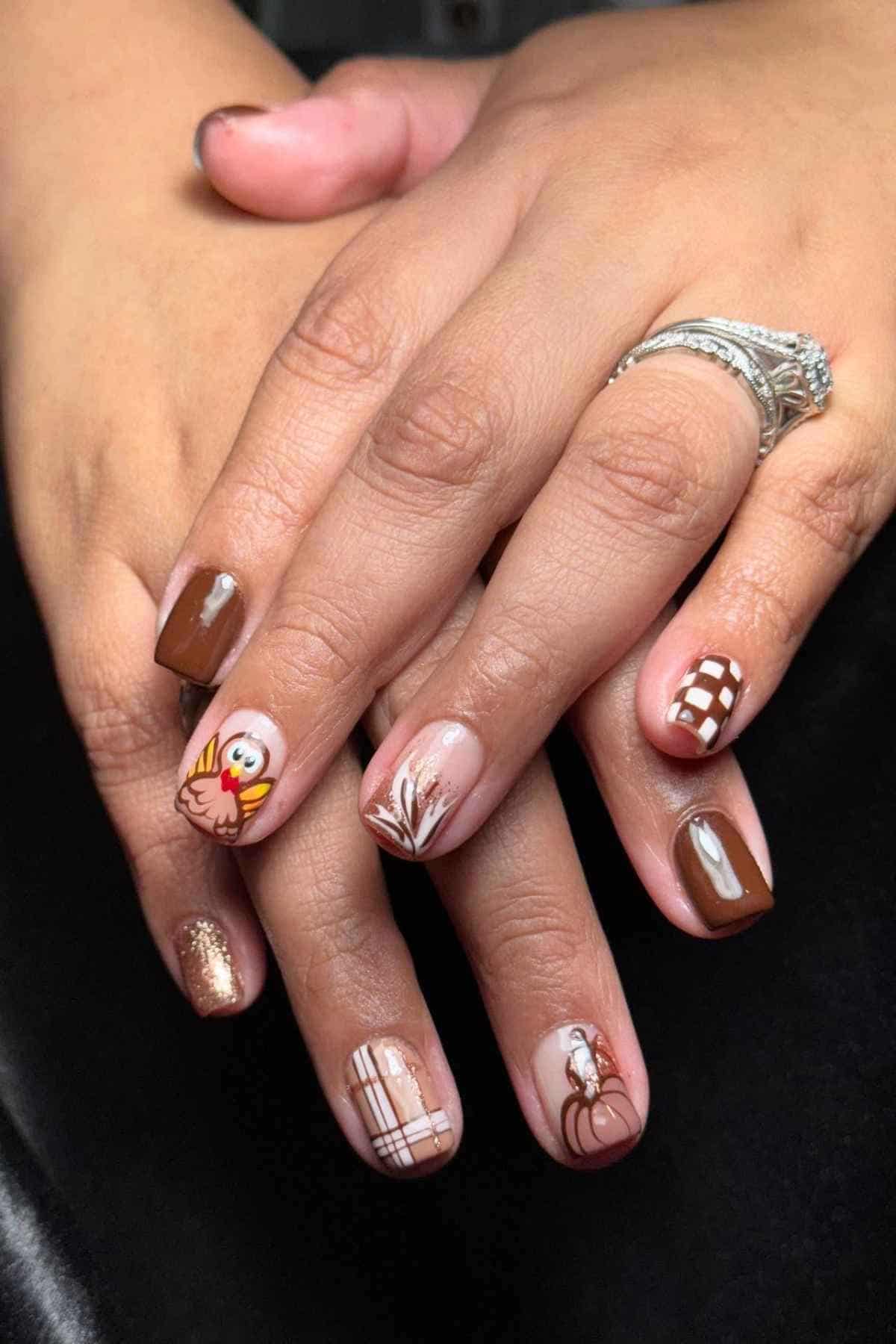 Hands with a Thanksgiving-themed manicure featuring brown, gold, and white nail art designs including a turkey, pumpkin, plaid, and checkerboard patterns.
