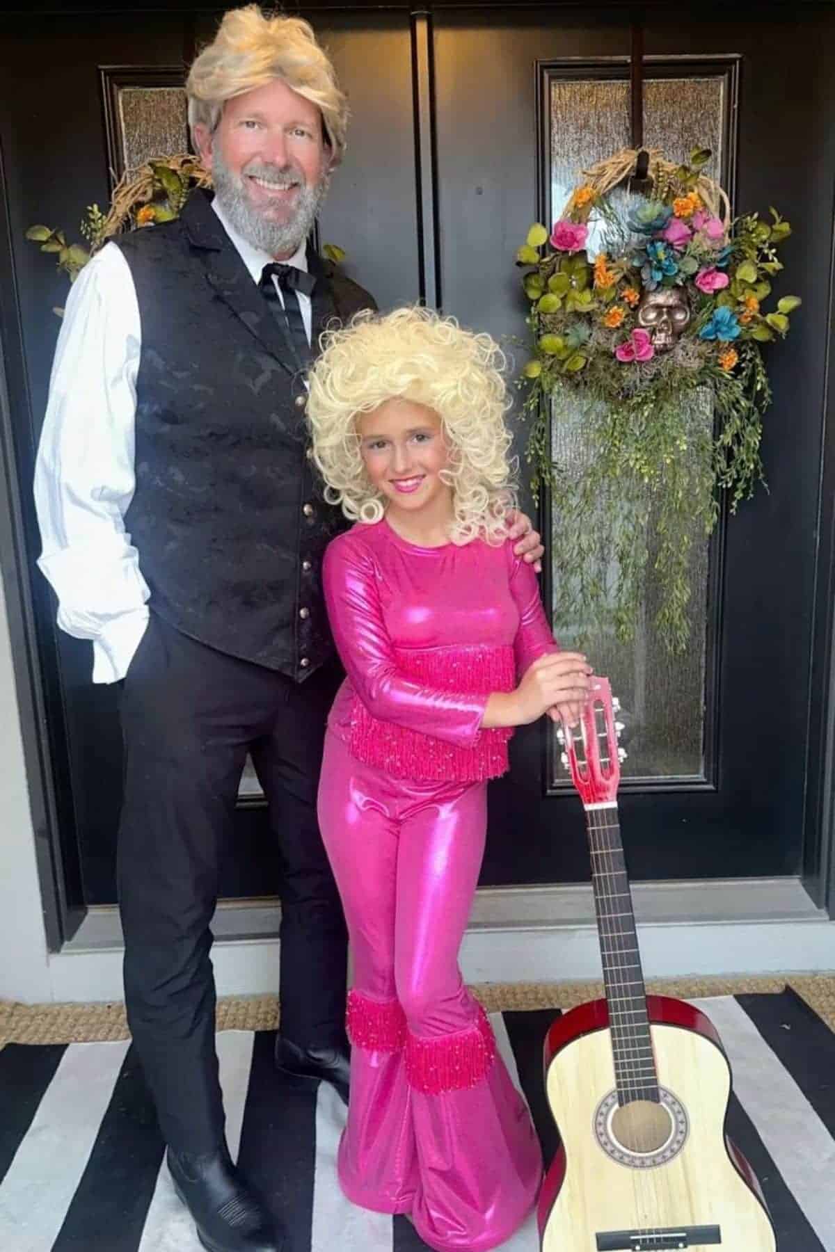 An adult and a child in blonde wigs pose in front of a door; the child wears a shiny pink outfit and stands next to a guitar.
