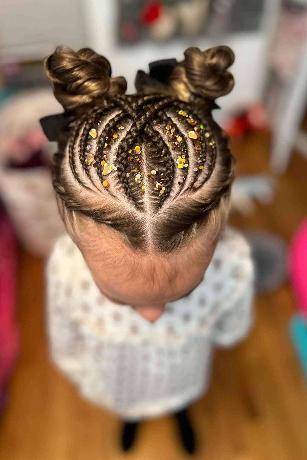 A child’s hair styled in symmetrical cornrows leading to two buns, decorated with gold glitter.