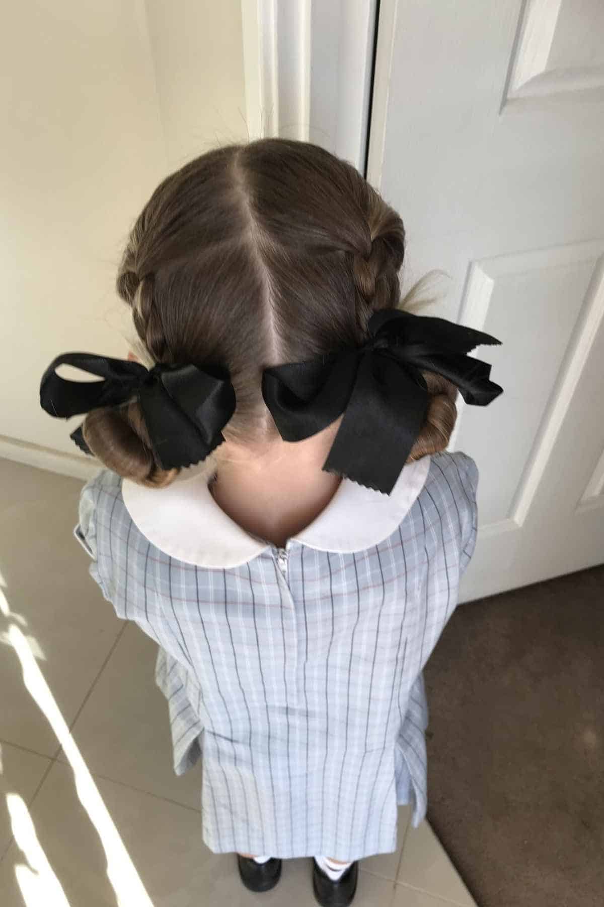 A girl from behind wearing a plaid school dress and hair styled in two buns with large black bows.