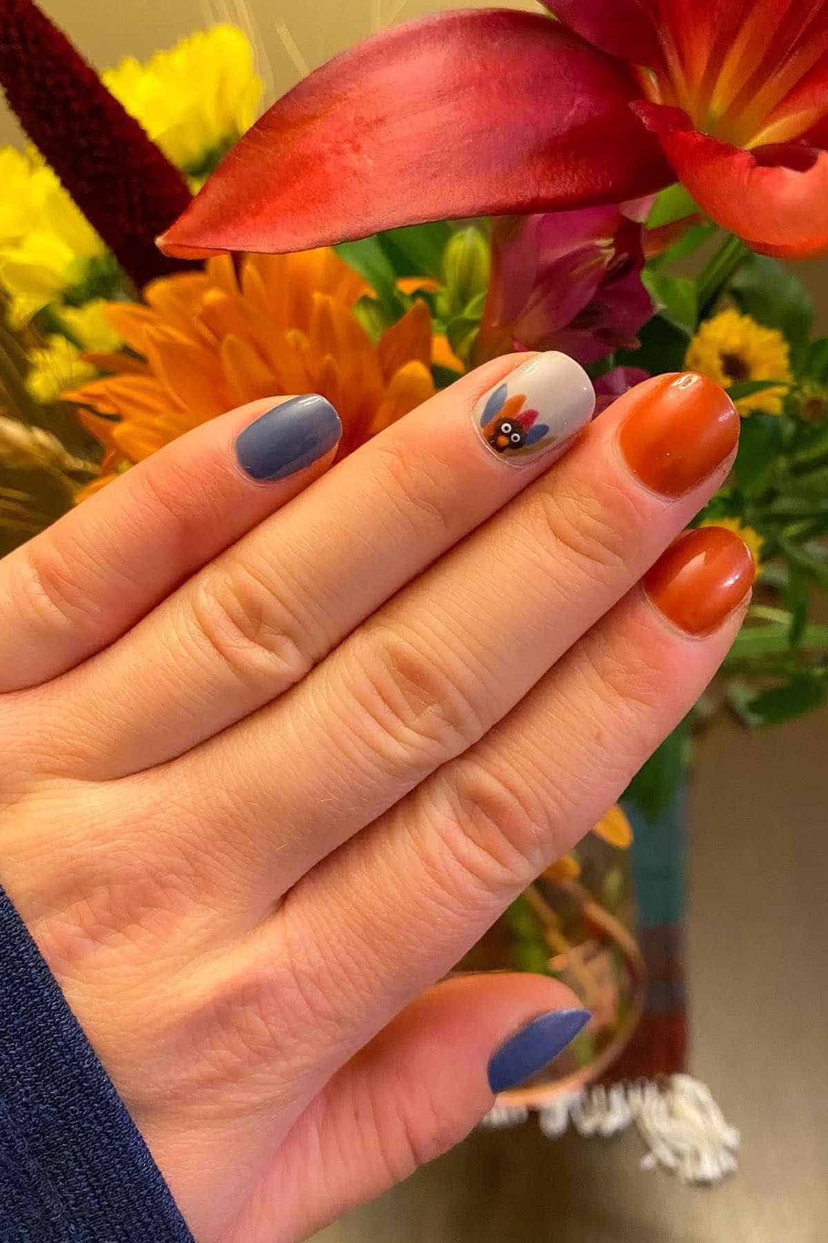 Hand with fall-themed nail polish in blue, orange, and a turkey-patterned accent nail, held in front of a bouquet of flowers.
