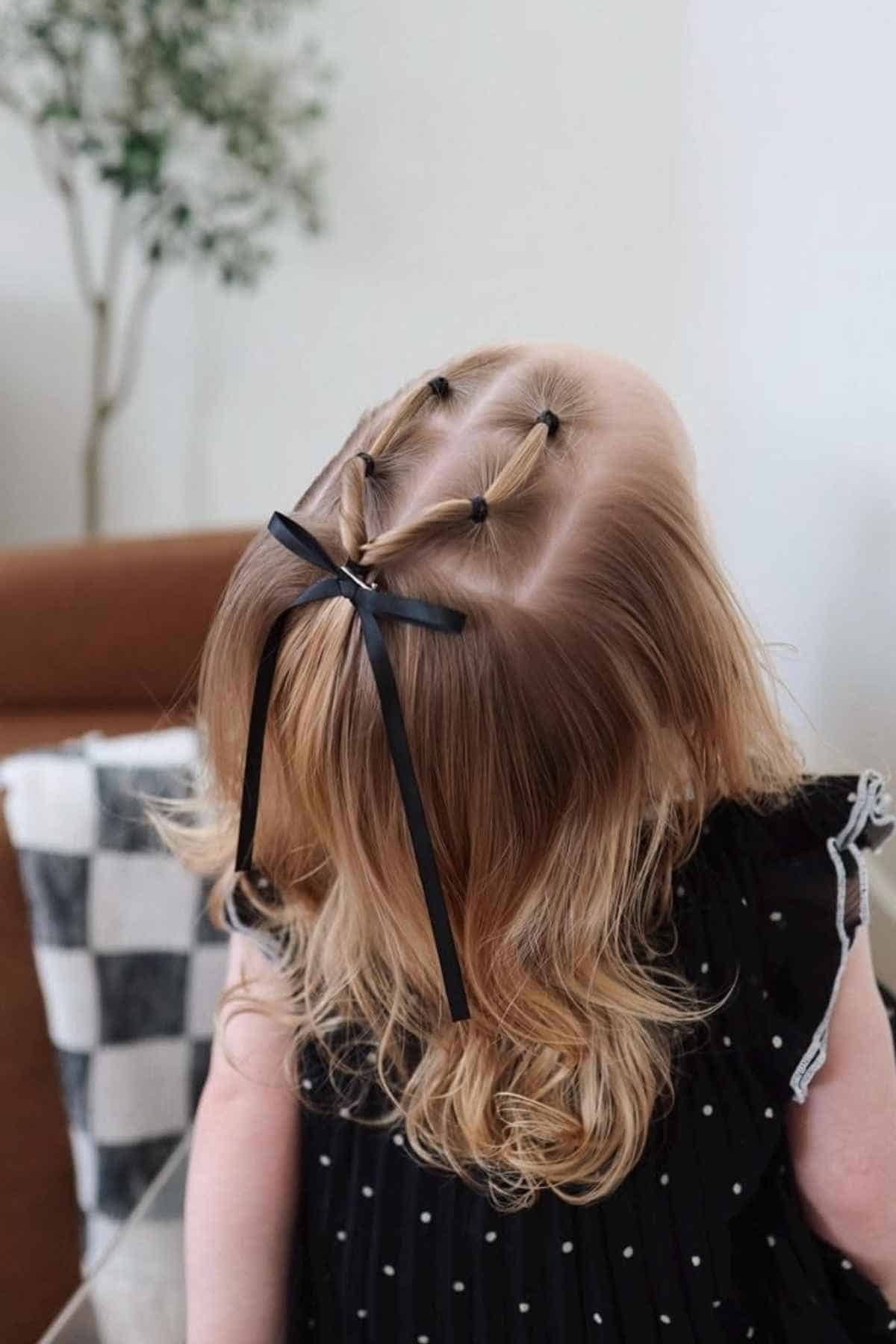A child with blonde hair styled in two small ponytails pulled back into a single larger ponytail secured with a black ribbon bow.