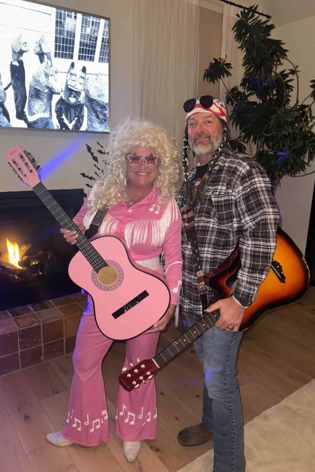 Two adults in costumes pose with guitars indoors; one wears a blonde wig and pink outfit, the other wears a headband and plaid shirt. A fireplace and TV are in the background.