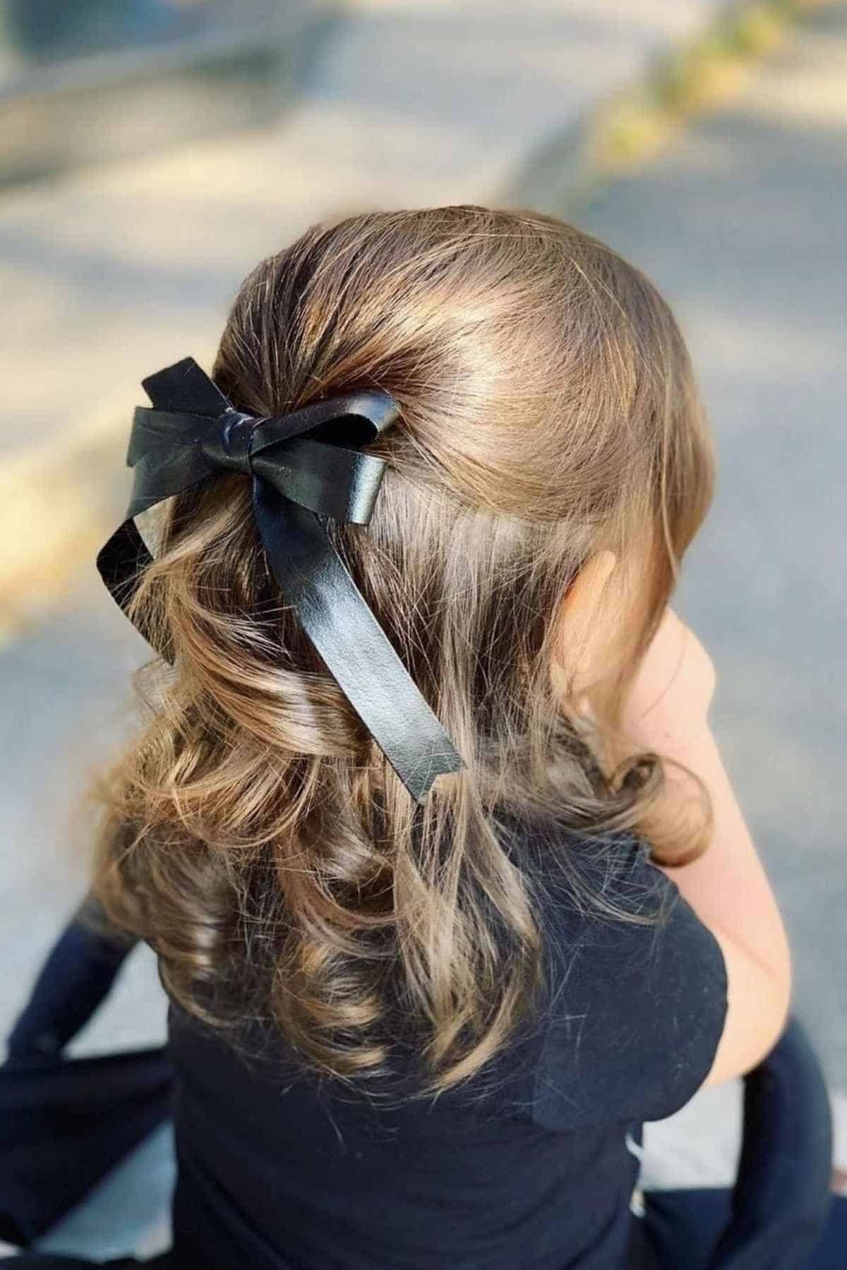 A child with wavy brown hair pulled back with a black ribbon bow.