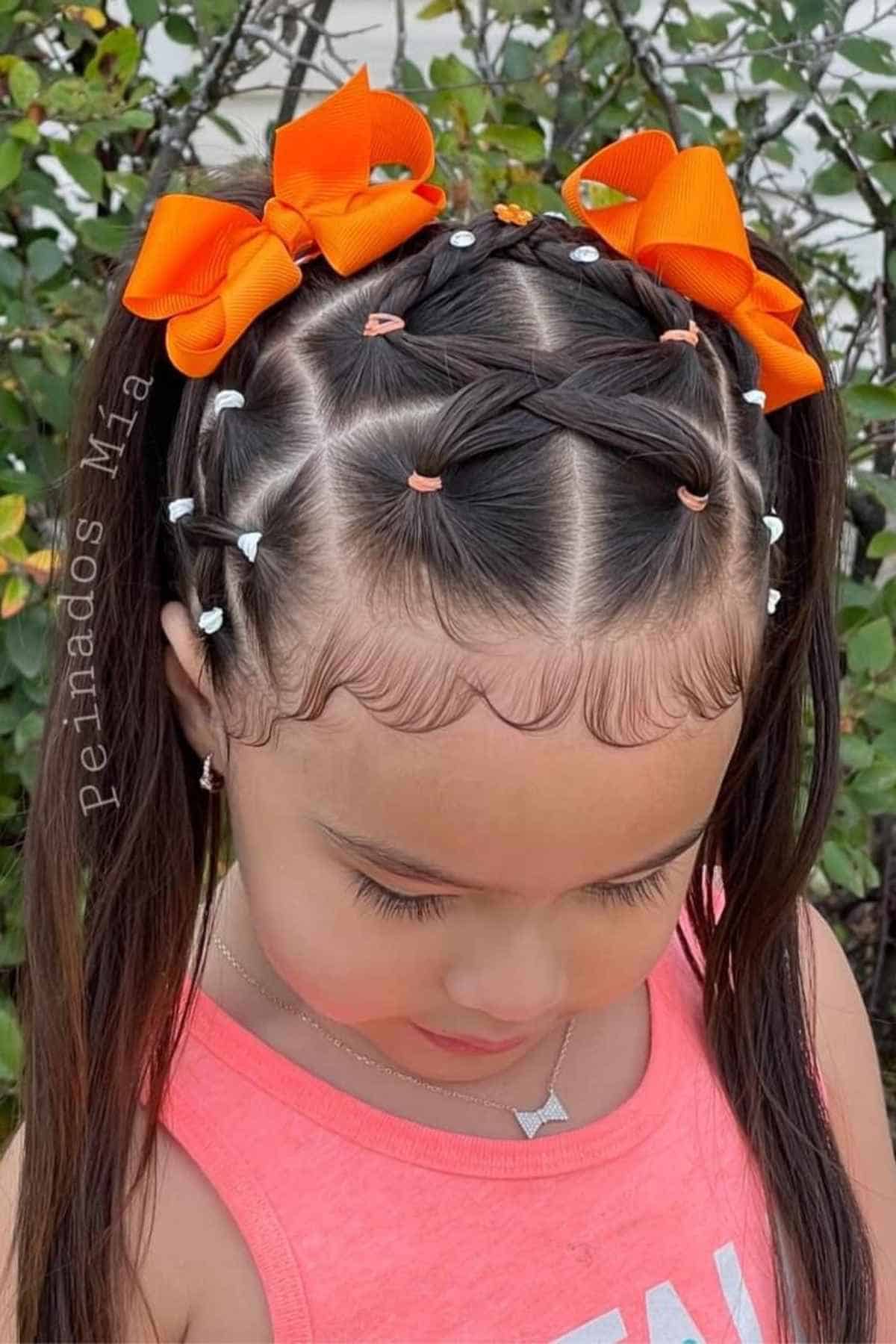 A young girl with a complex hairstyle consisting of braided sections, small colorful hair elastics, and two large orange hair bows.