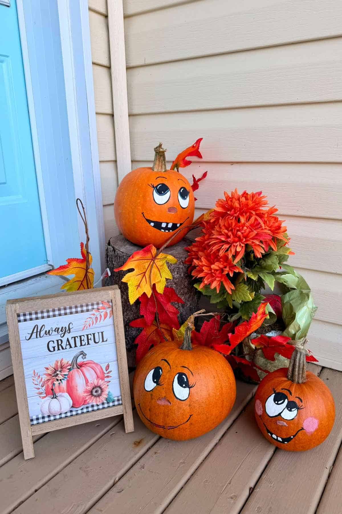 Three painted pumpkins with faces, artificial fall flowers, and a small sign that reads “Always Grateful” on a porch.