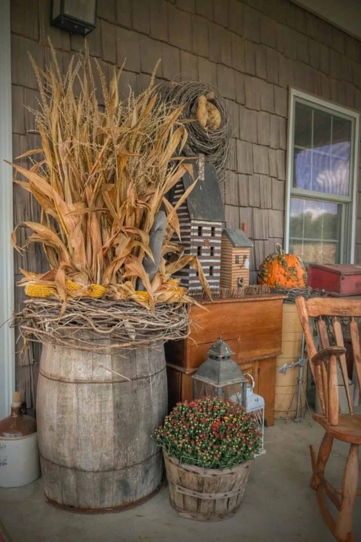 Fall porch decor featuring dried corn stalks in a wooden barrel, a small wooden house, a pumpkin, and a basket of potted mums.