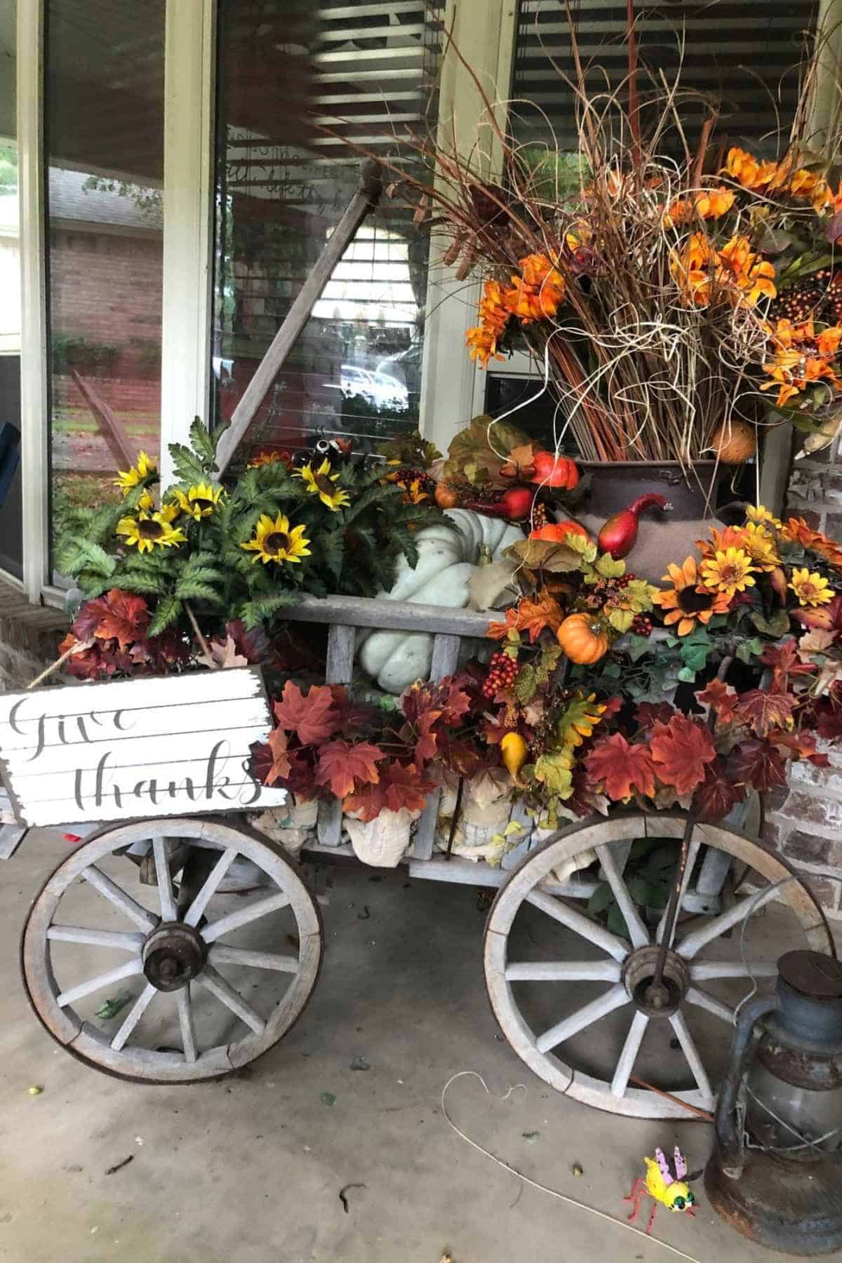 A wooden garden cart decorated with autumn foliage, sunflowers, pumpkins, and a sign that reads “Give thanks.”
