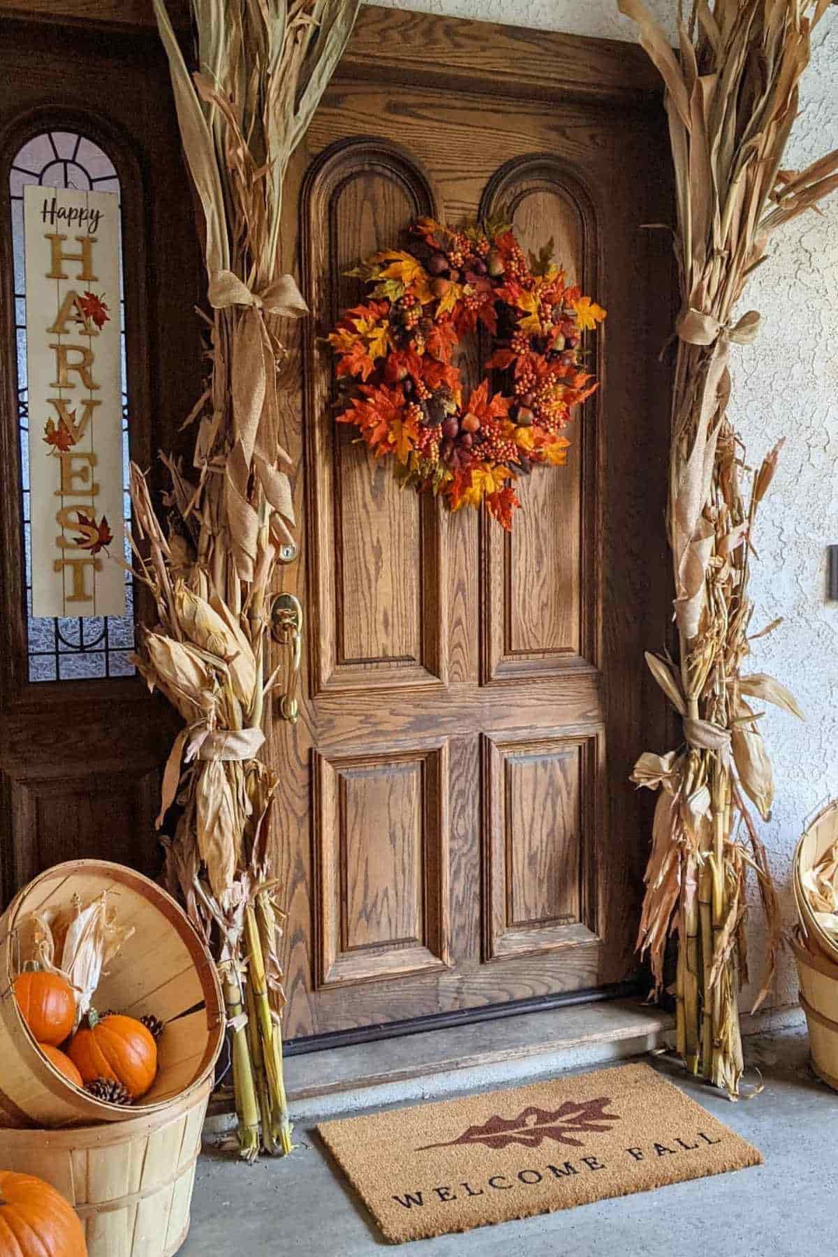 Front door decorated for autumn with a fall-colored wreath, stalks of corn on the doorframe, a “Happy Harvest” sign, and a “Welcome Fall” doormat with pumpkins in a basket.