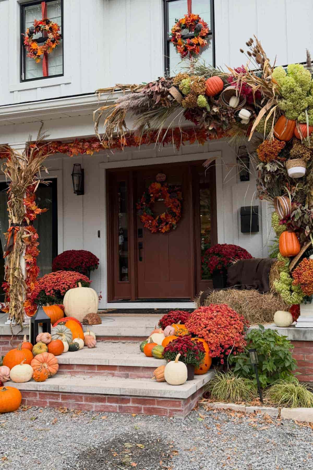 Front entrance of a house decorated for autumn with pumpkins, orange and red mums, dried corn stalks, and wreaths.