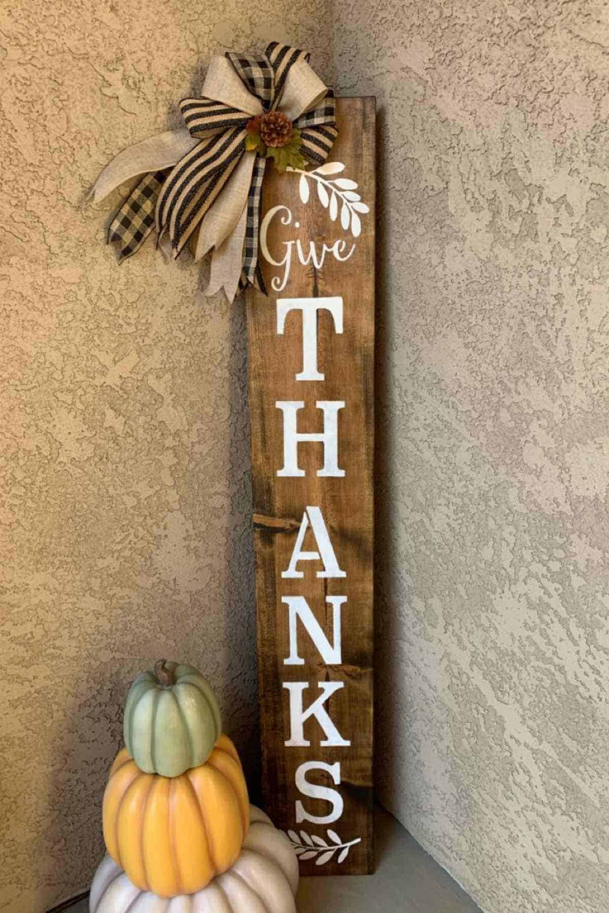 Tall wooden porch sign reading “Give THANKS” in white letters, decorated with a plaid bow and a stack of three small decorative pumpkins.