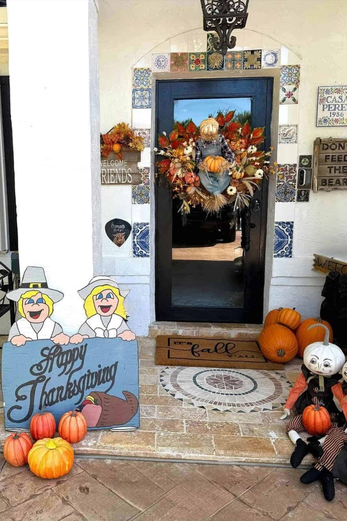 Front porch decorated for fall with a scarecrow wreath on the door, pumpkins, and a “Happy Thanksgiving” sign featuring two pilgrims.