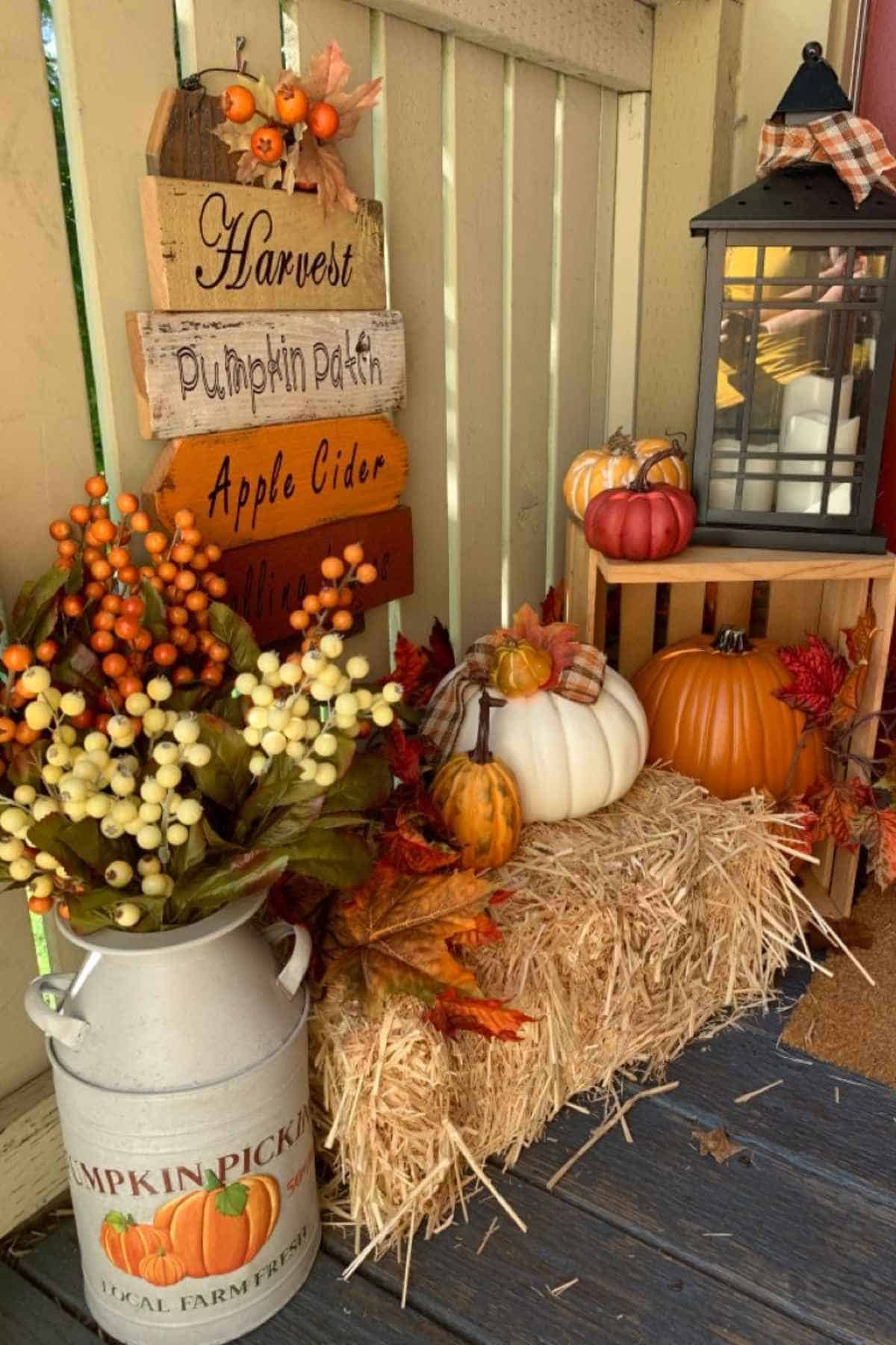 Autumn porch decor featuring a “Harvest” sign, a milk can with artificial berries, a straw bale, assorted pumpkins, and a lantern.