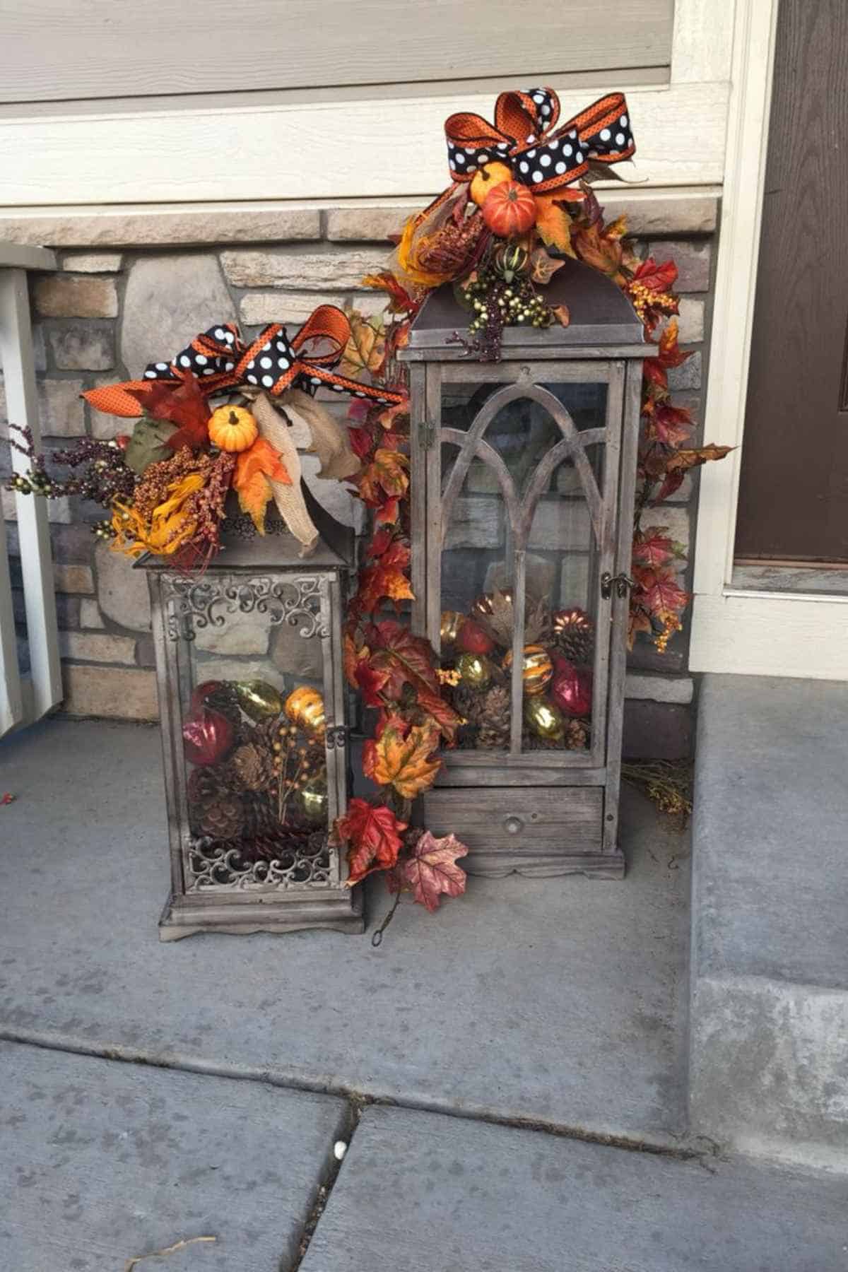 Two wooden lanterns decorated with autumn leaves, ribbons, and pinecones, sitting on a porch.