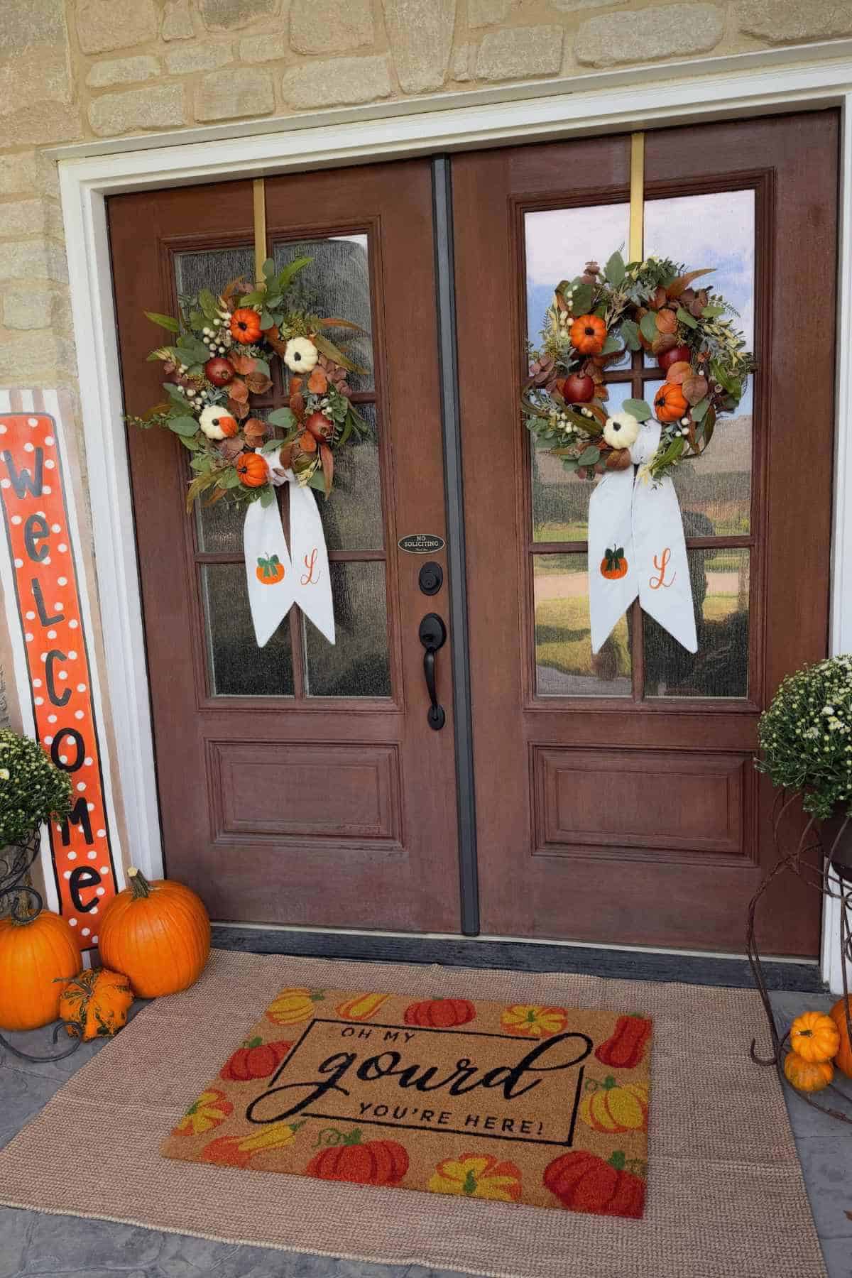Double front doors decorated with autumn wreaths, pumpkins, and a doormat that reads “Oh my gourd you’re here!”.