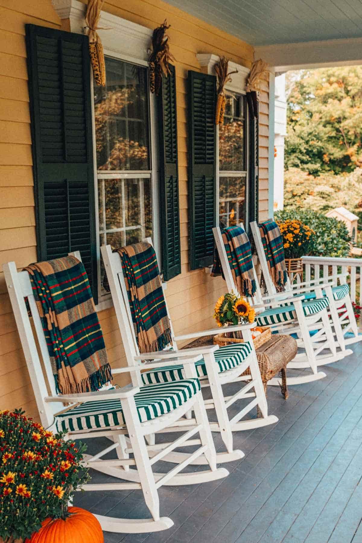 Two white rocking chairs with green-striped cushions and plaid blankets on a porch, separated by a small table with a sunflower bouquet.