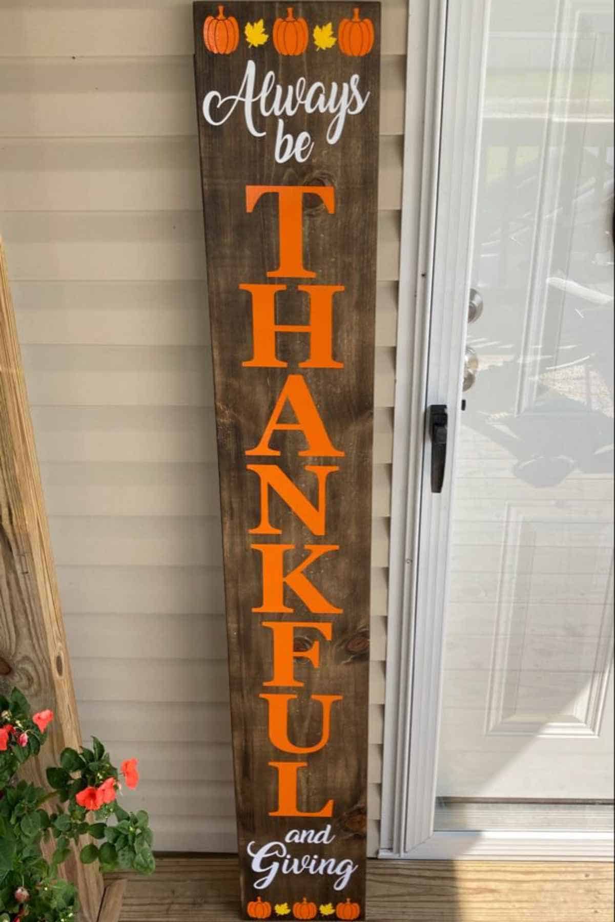 A vertical wooden porch sign that reads “Always be THANKFUL and Giving” in orange lettering, decorated with pumpkins at the top and bottom.