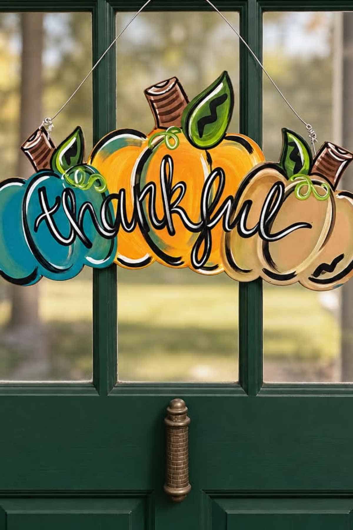 Hanging door sign featuring three pumpkins in blue, yellow, and tan, with the word “thankful” written in white cursive script across them.