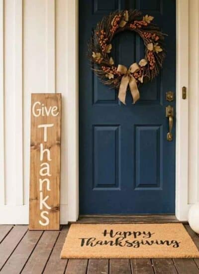 A front porch with Thanksgiving decor ideas, such as wreath, wooden sign, mat, pumpkins, and mums.