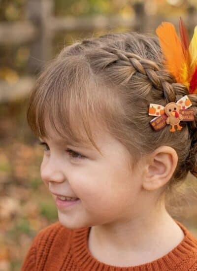 A young girl with a braided hairstyle decorated with colorful feathers and a small turkey-themed hair clip.