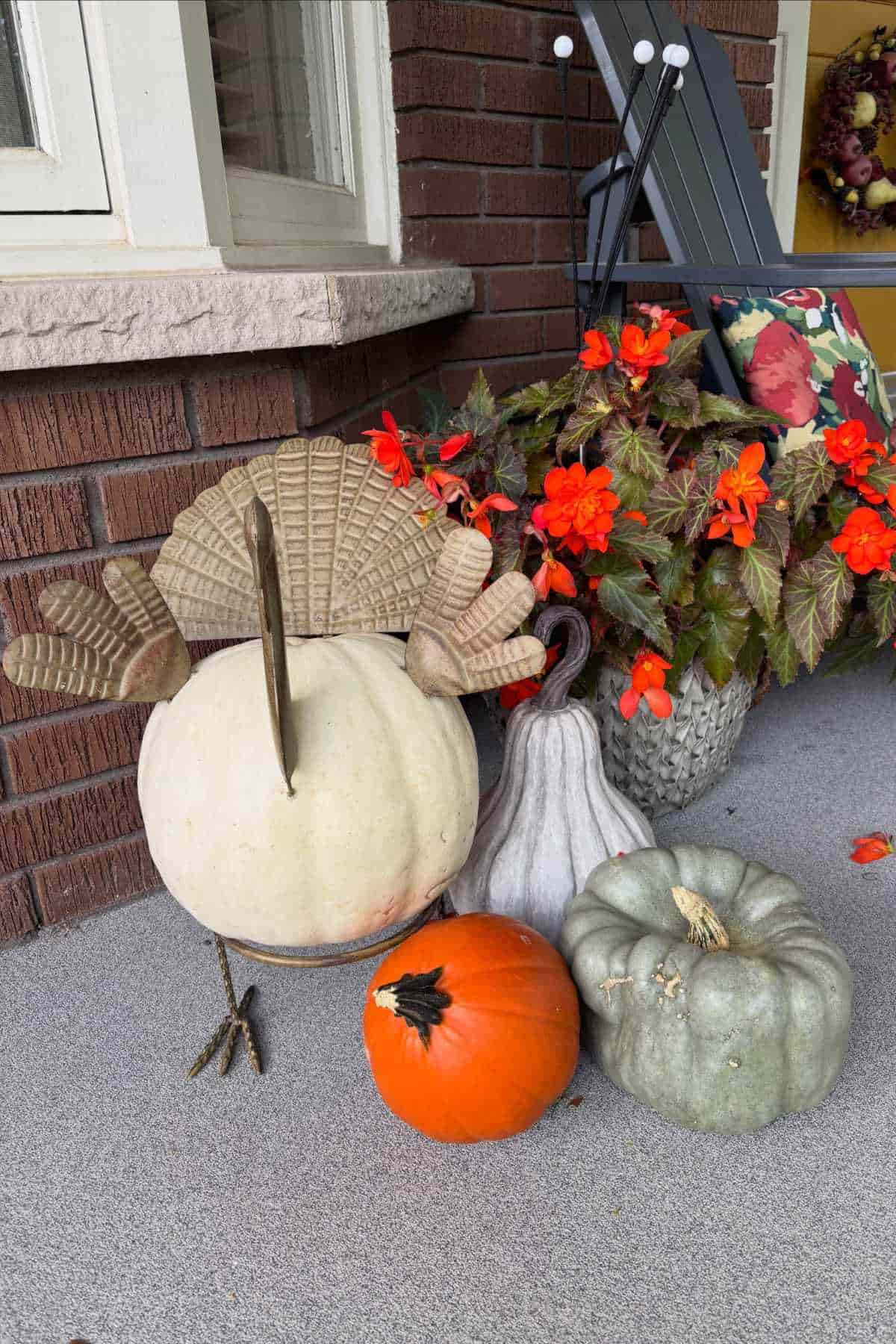 A white pumpkin decorated as a turkey with a metal fan tail and feet, sitting on a porch with three other pumpkins and a pot of orange flowers.
